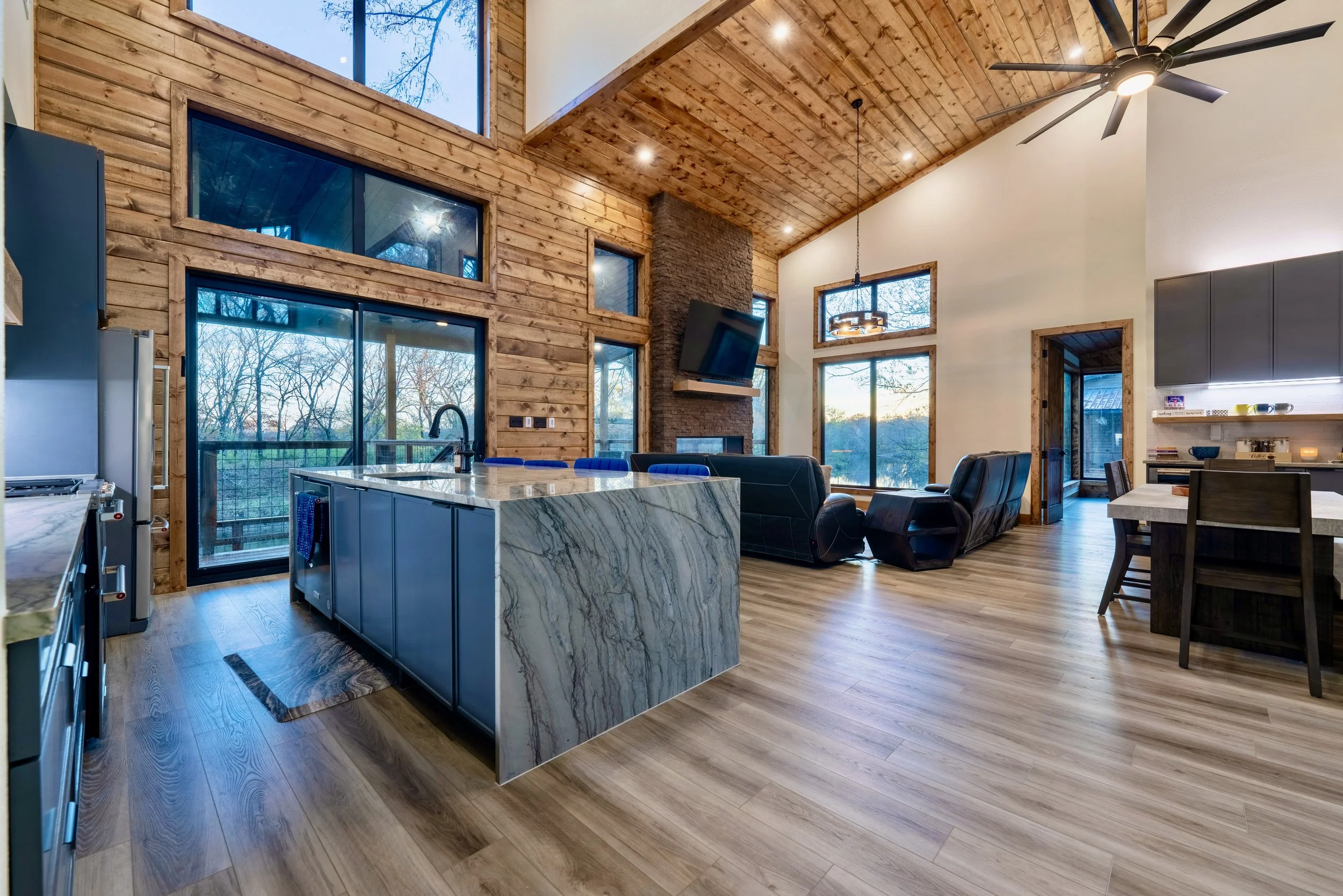 Open-concept living area with high wooden ceiling, large windows, stone fireplace, modern kitchen with marble island, black leather chairs, and a dining table.