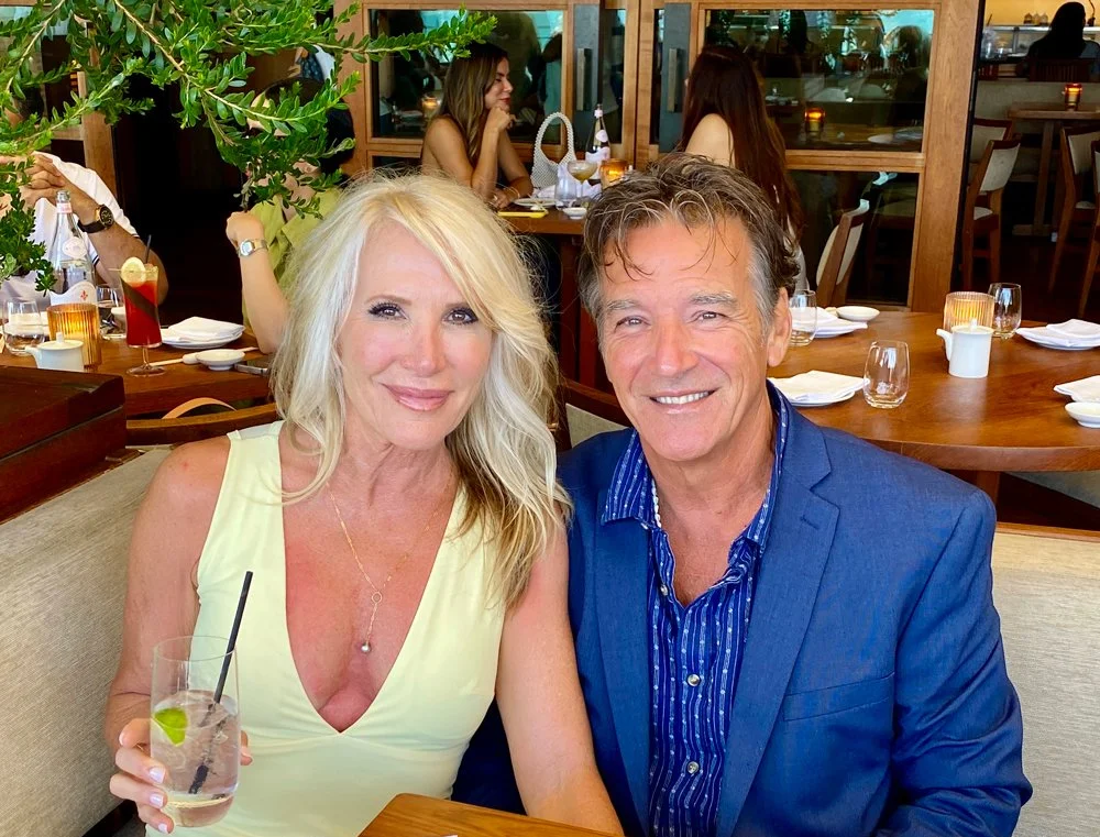A smiling blonde woman in a yellow dress holding a glass with ice, lime, and a straw, sitting next to a smiling man in a blue blazer and striped shirt at a restaurant.