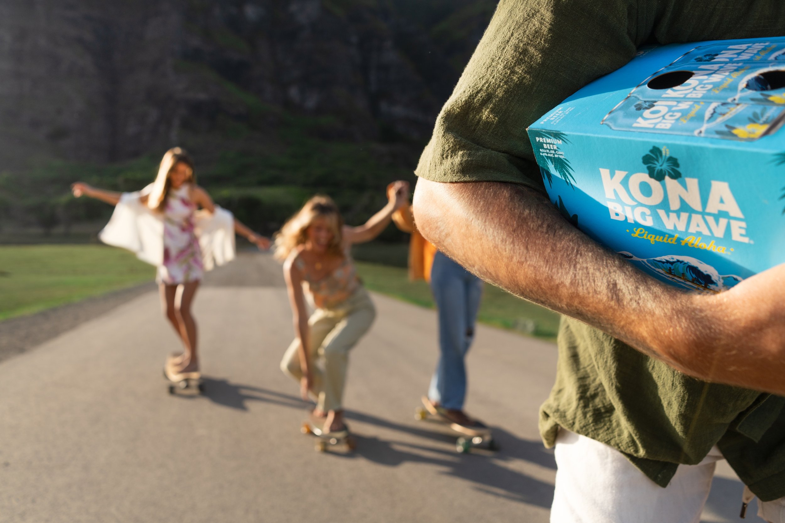 Person holding a box of Kona Big Wave beer in front of three children roller skating outdoors with a mountainous landscape in the background.