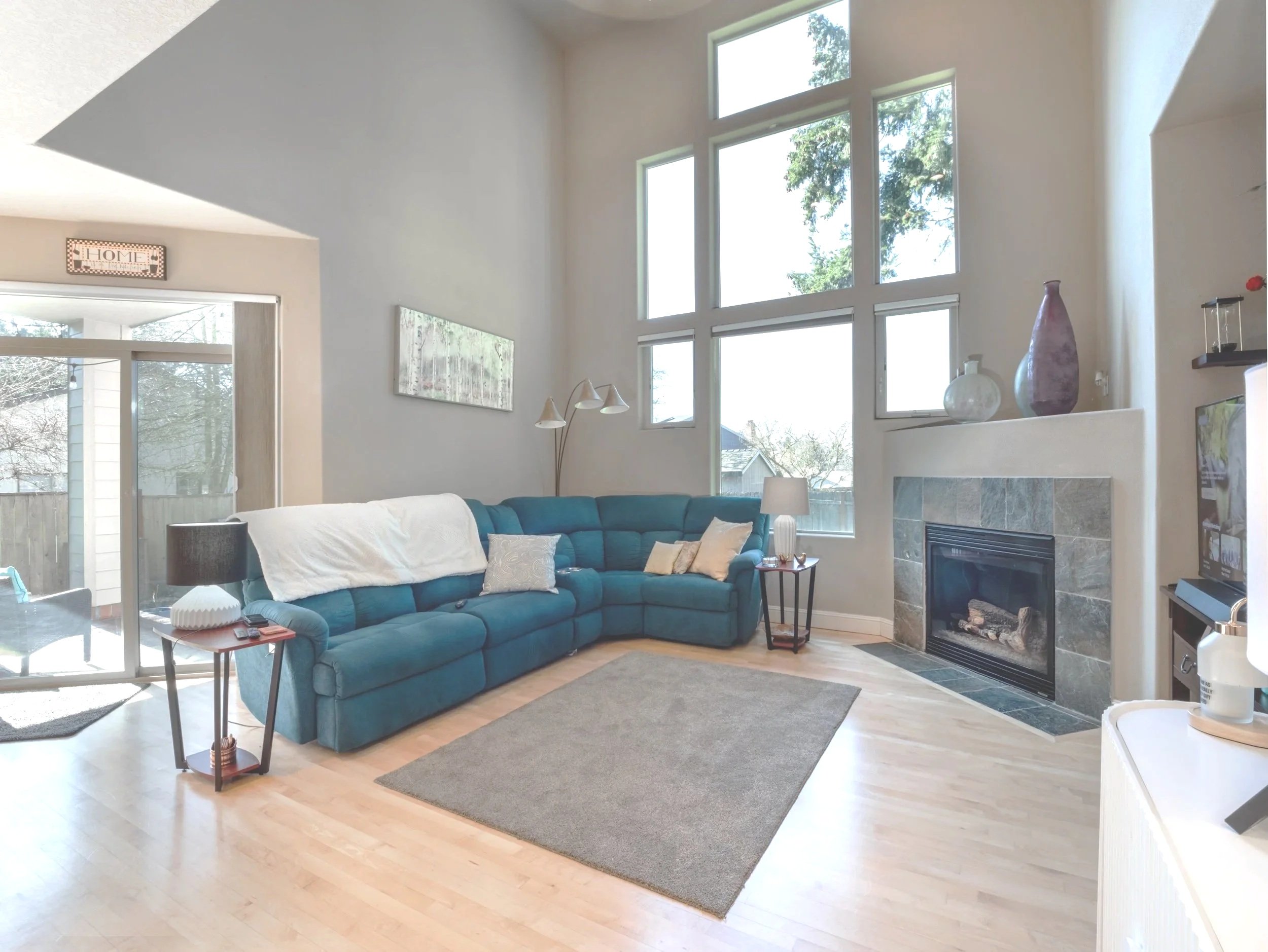 Living room with tall windows, teal sectional sofa, fireplace with stone surround, wooden floor, side tables with lamps, and decorative vases on mantle.