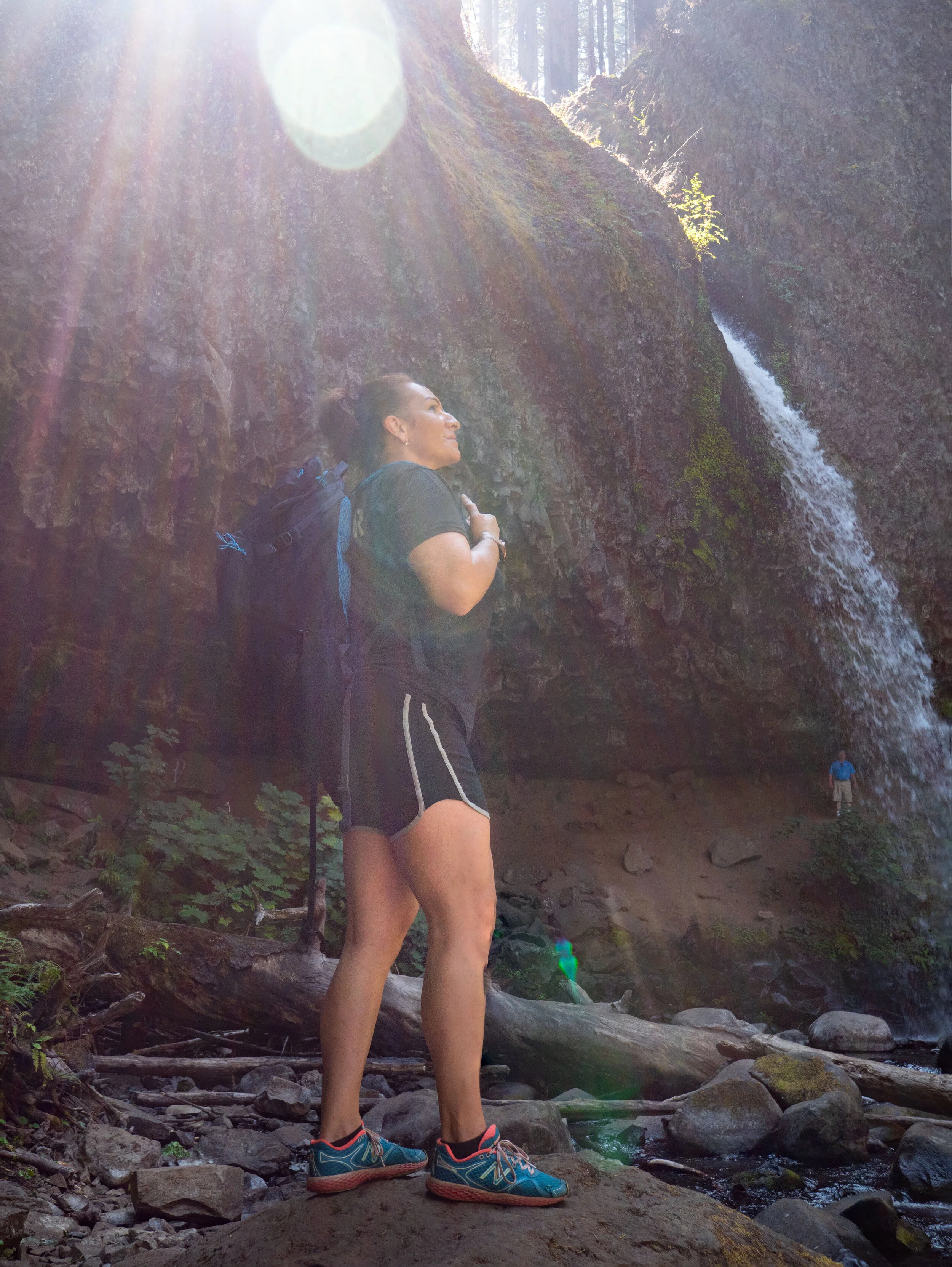 A woman standing on rocks near a waterfall in a forest, with sunlight shining through trees.