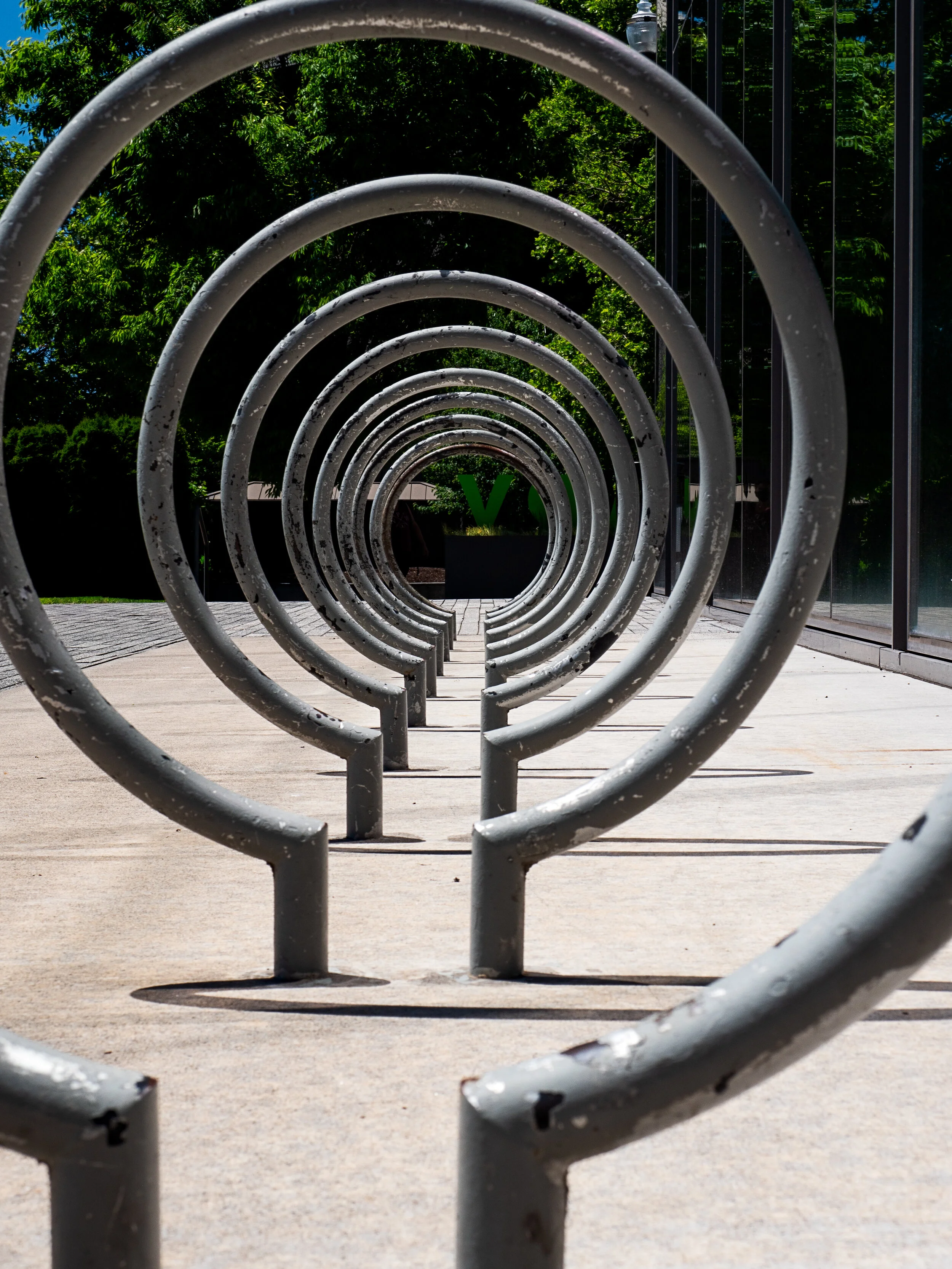 A row of metal bike racks forming a spiral tunnel on a sidewalk with greenery and a glass building in the background.