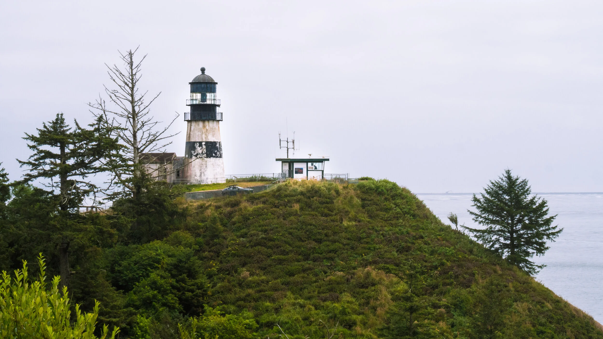 A lighthouse on a grassy hill with trees, overlooking the ocean on a cloudy day.