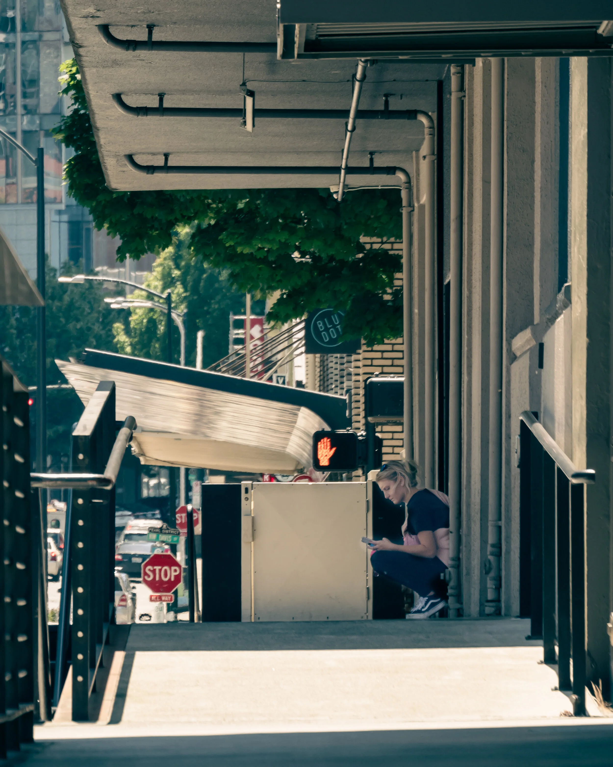 City sidewalk scene with a woman sitting on a step, looking at her phone, pedestrian crossing signal showing a red hand, stop signs, trees, and buildings in the background.