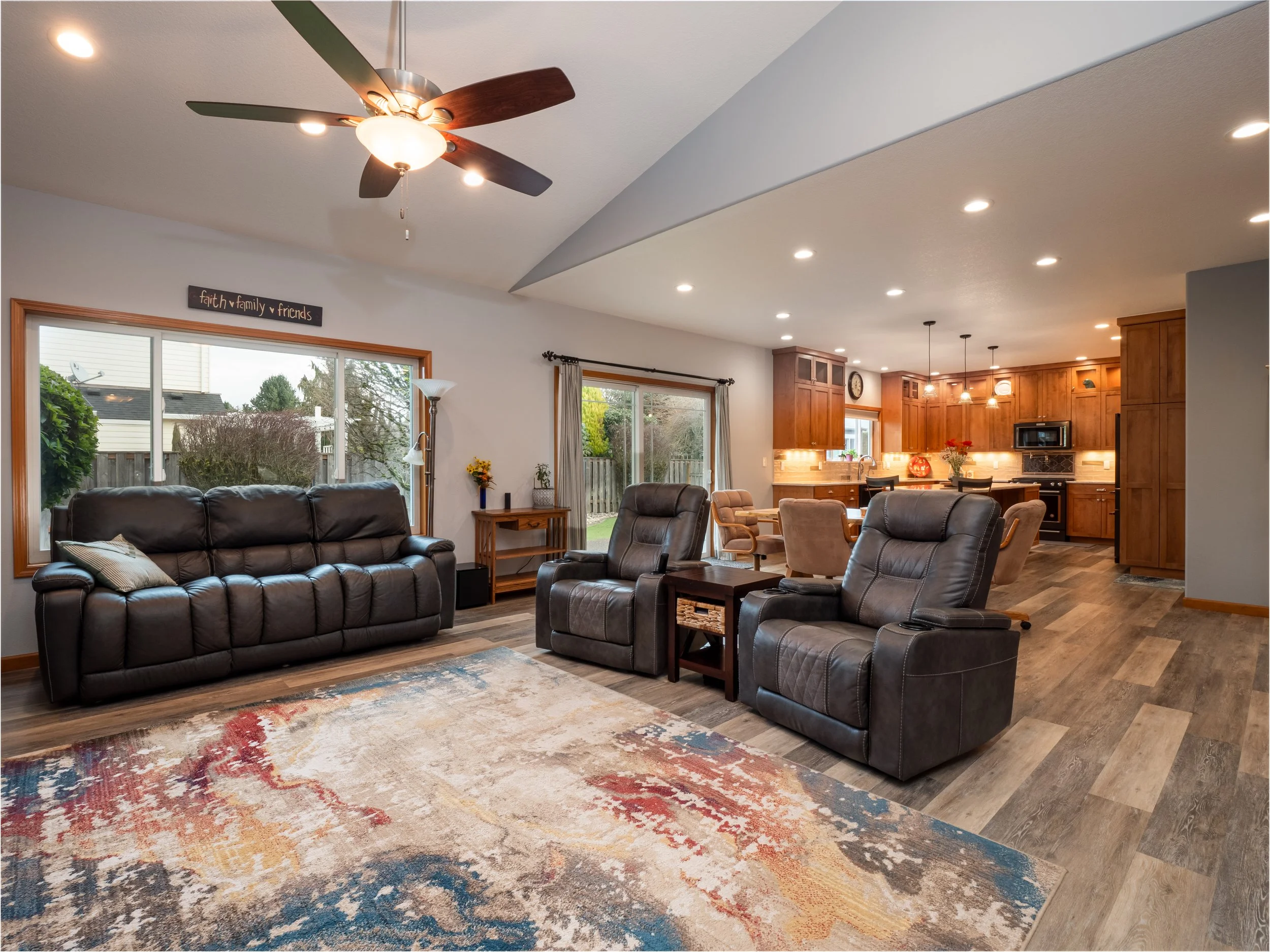 Open-plan living room with leather sofa and armchairs, rug, ceiling fan, standing lamp, large windows, view of backyard, connected to kitchen with wooden cabinets, dining table, and pendant lights.