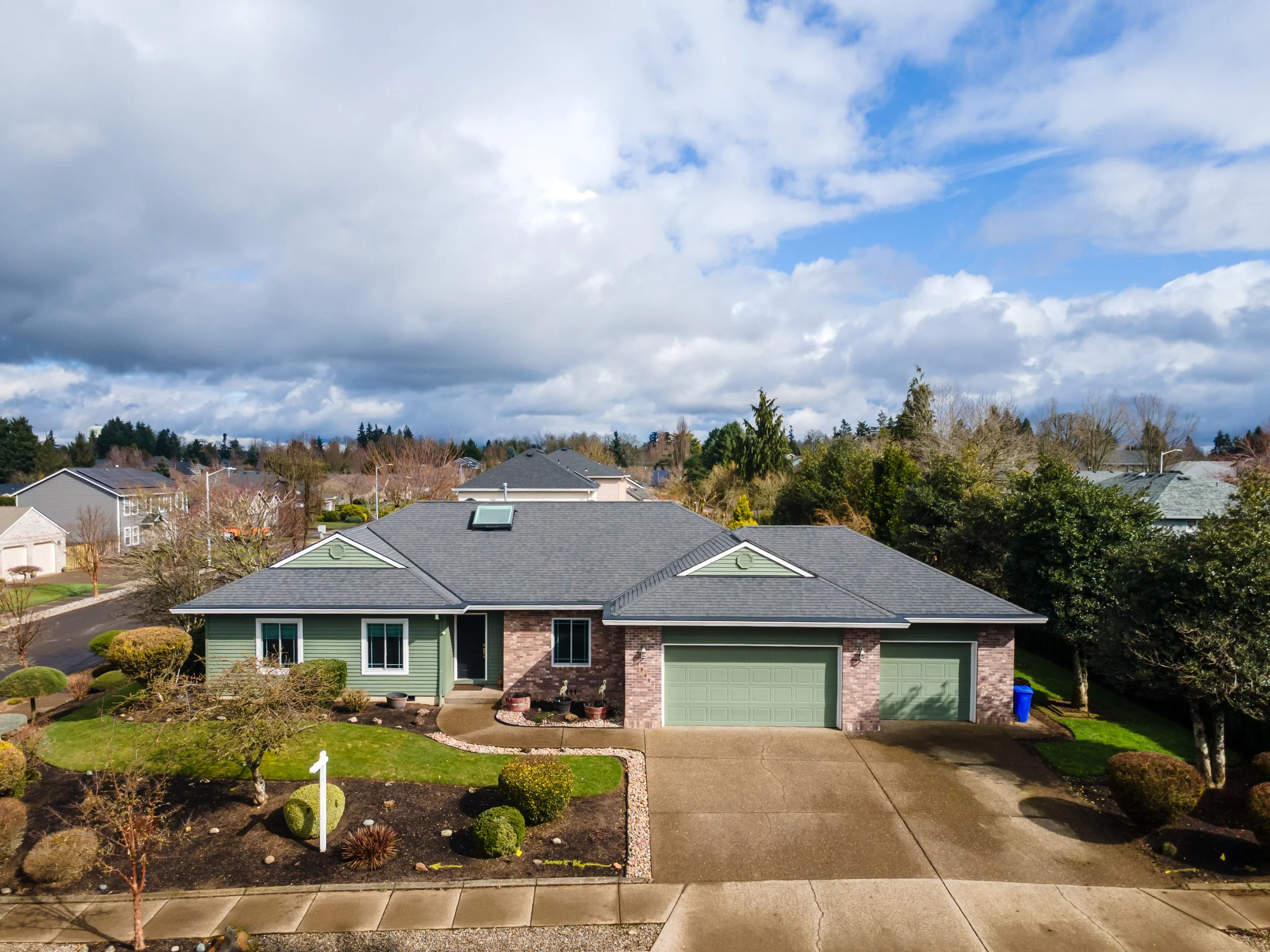 Front yard of a modern house with a gray roof, green siding, and a brick accent wall, surrounded by a manicured lawn and trees, under a partly cloudy sky.