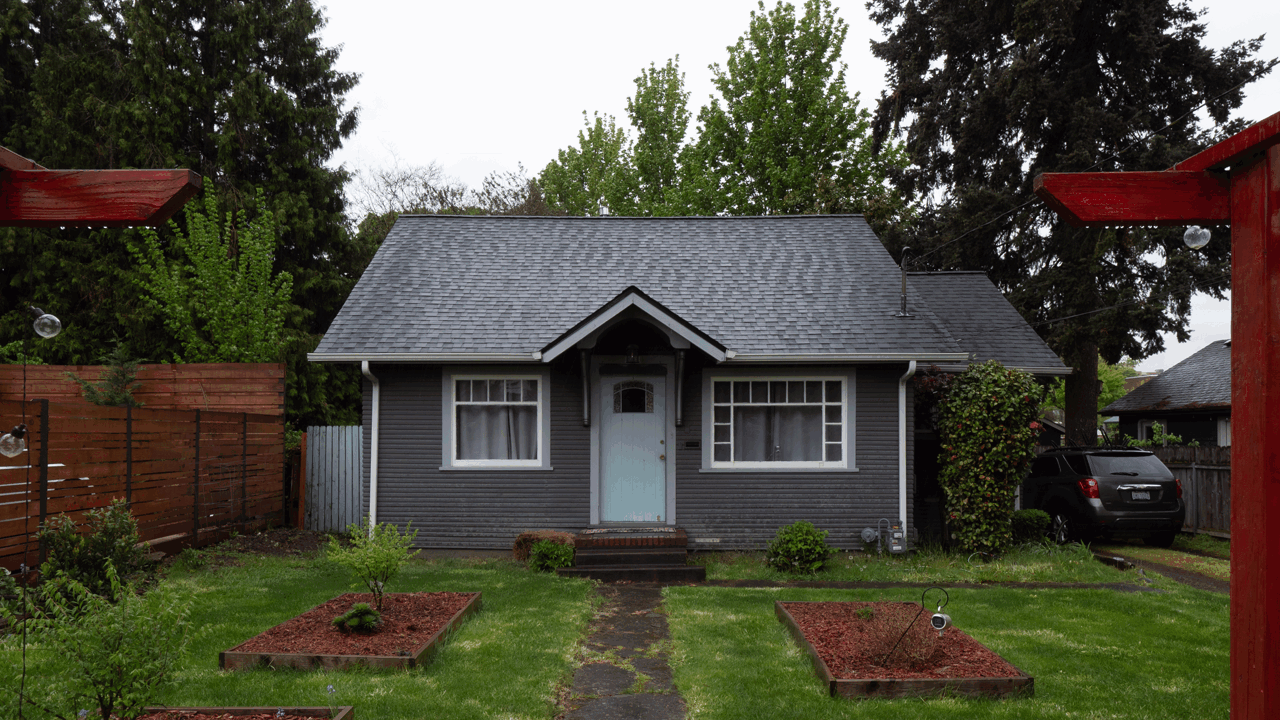 Front view of a small, gray house with white trim, a steep roof, and a front door with steps. The house is surrounded by a green lawn, trees, and fenced garden beds. There is a black car parked on the side.