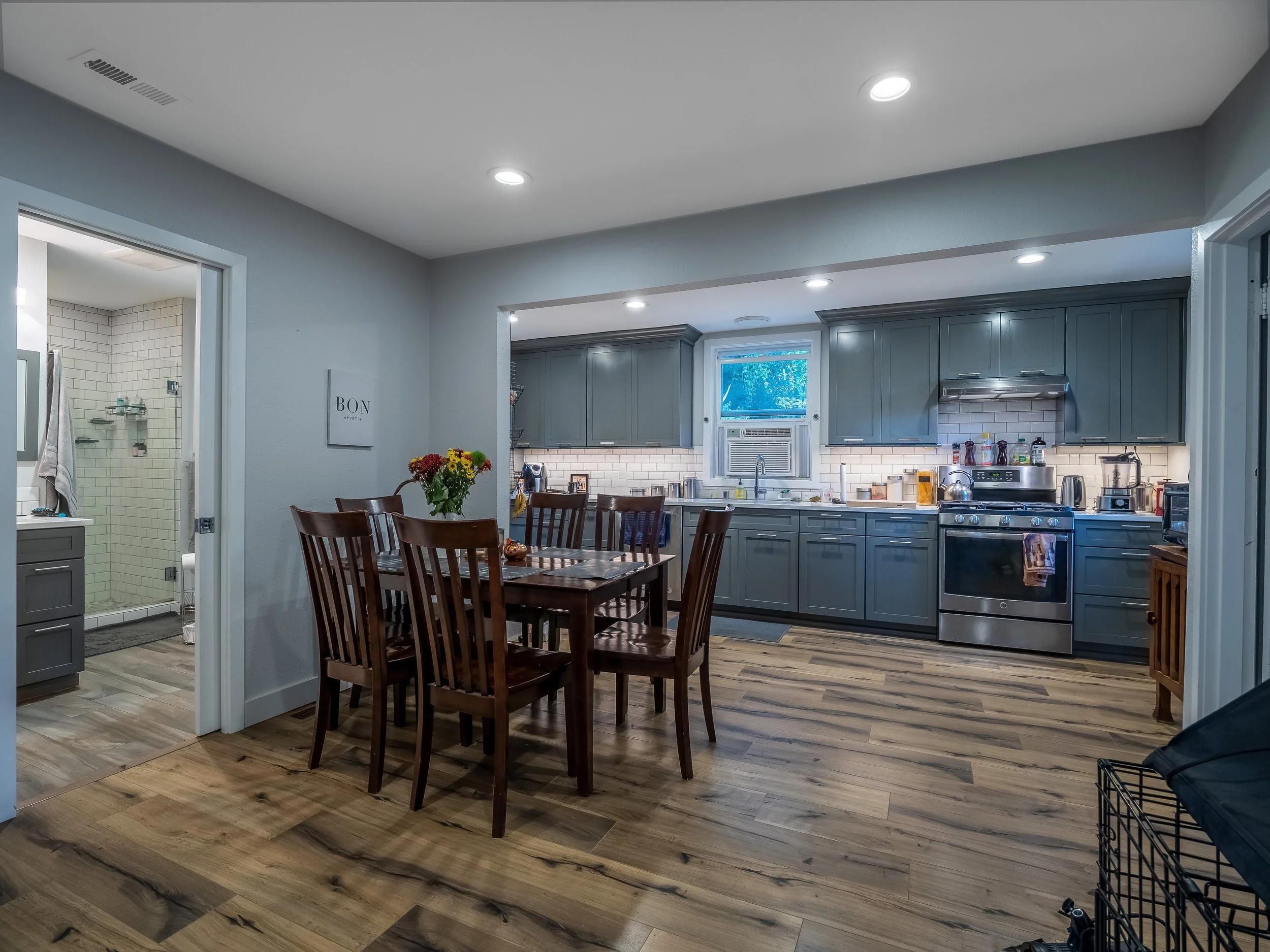 View of a modern kitchen with gray cabinets, stainless steel appliances, white subway tile backsplash, a window above the sink, and a wooden dining table with chairs. A closet with a bathroom is seen on the left.