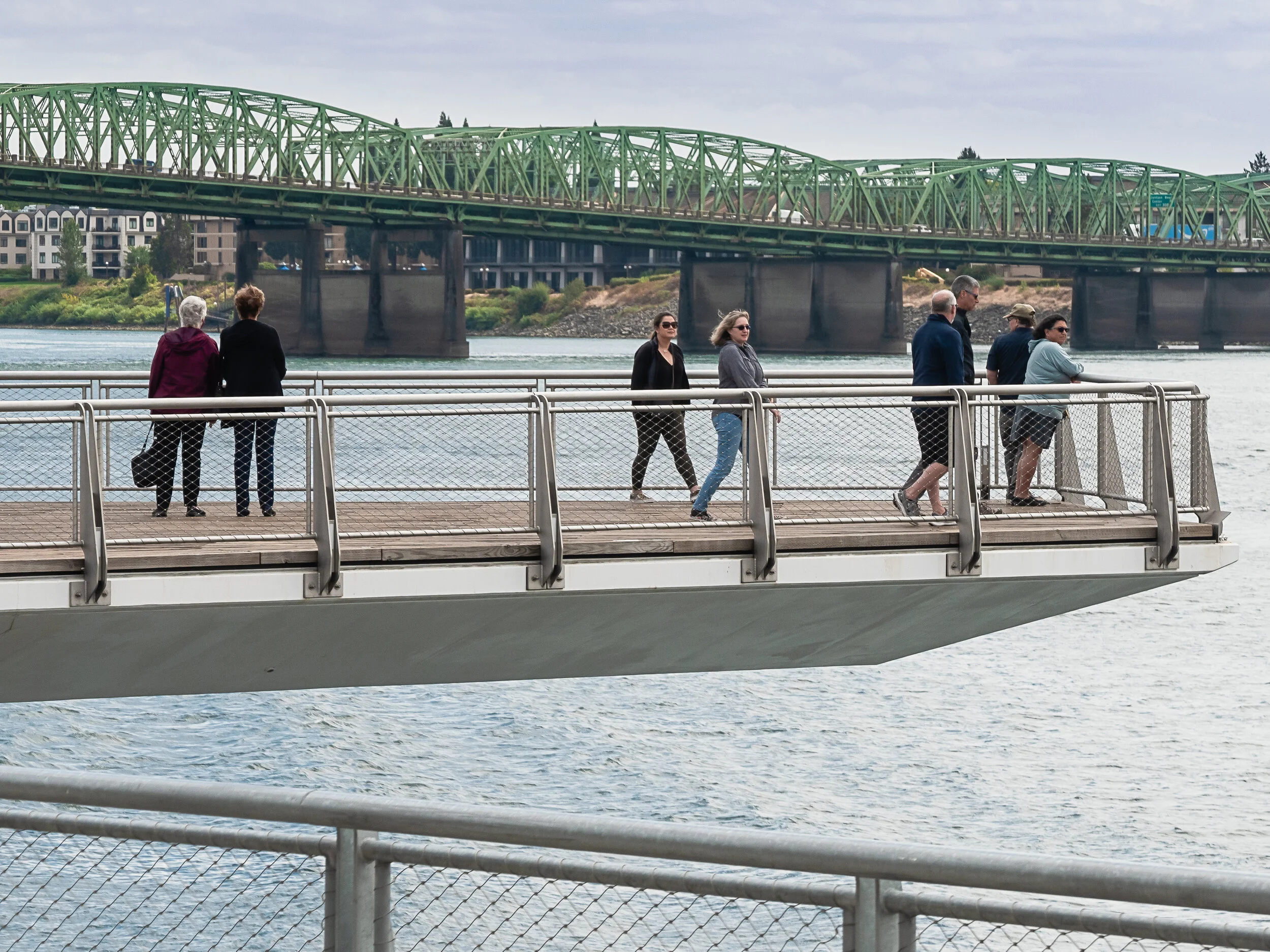 People walking and standing on a waterfront pier with a green bridge and city buildings in the background.