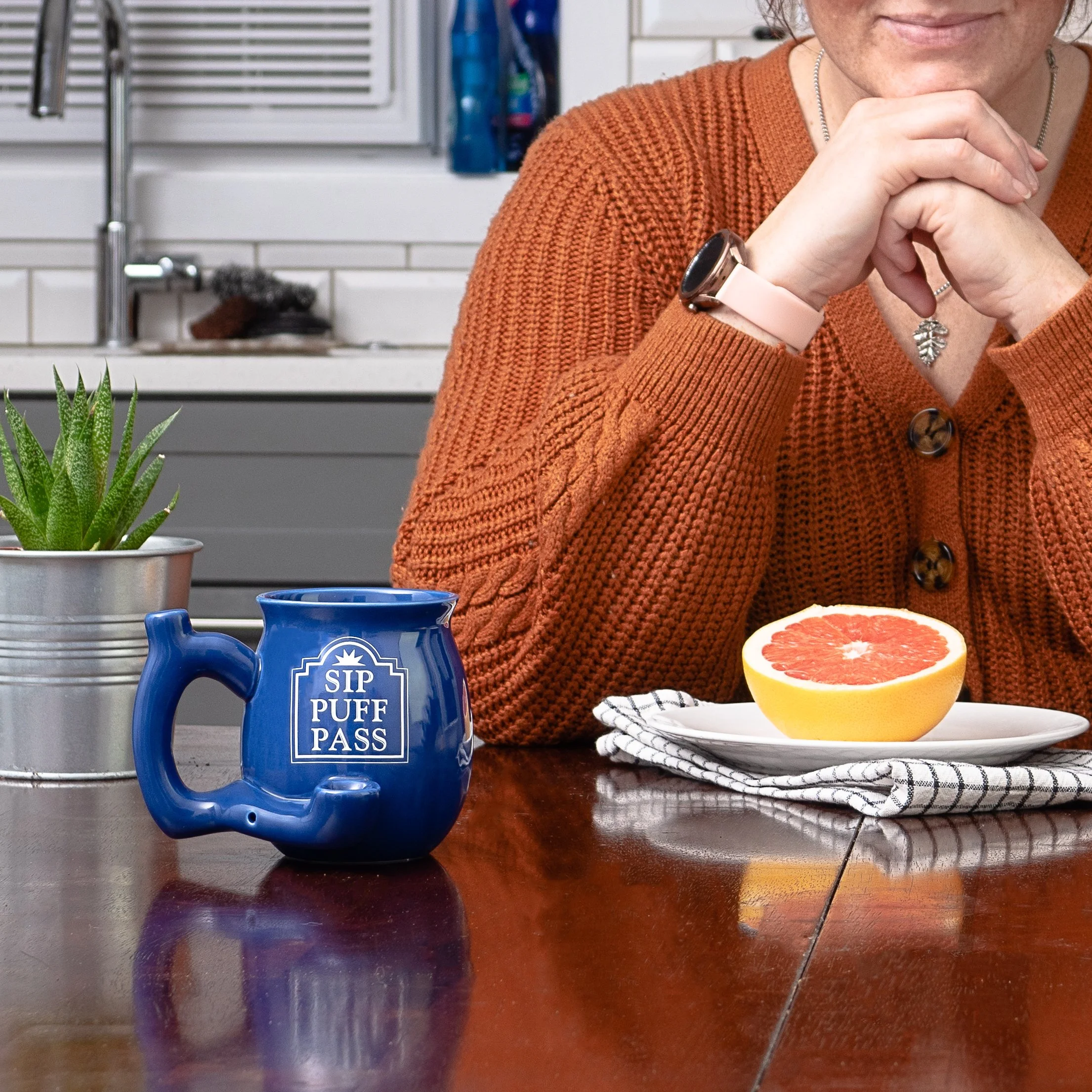 Part of a woman with a brown sweater, sitting at a wooden table with a half grapefruit, a potted plant, and a blue mug with white text that reads 'SIP PUFF PASS'.