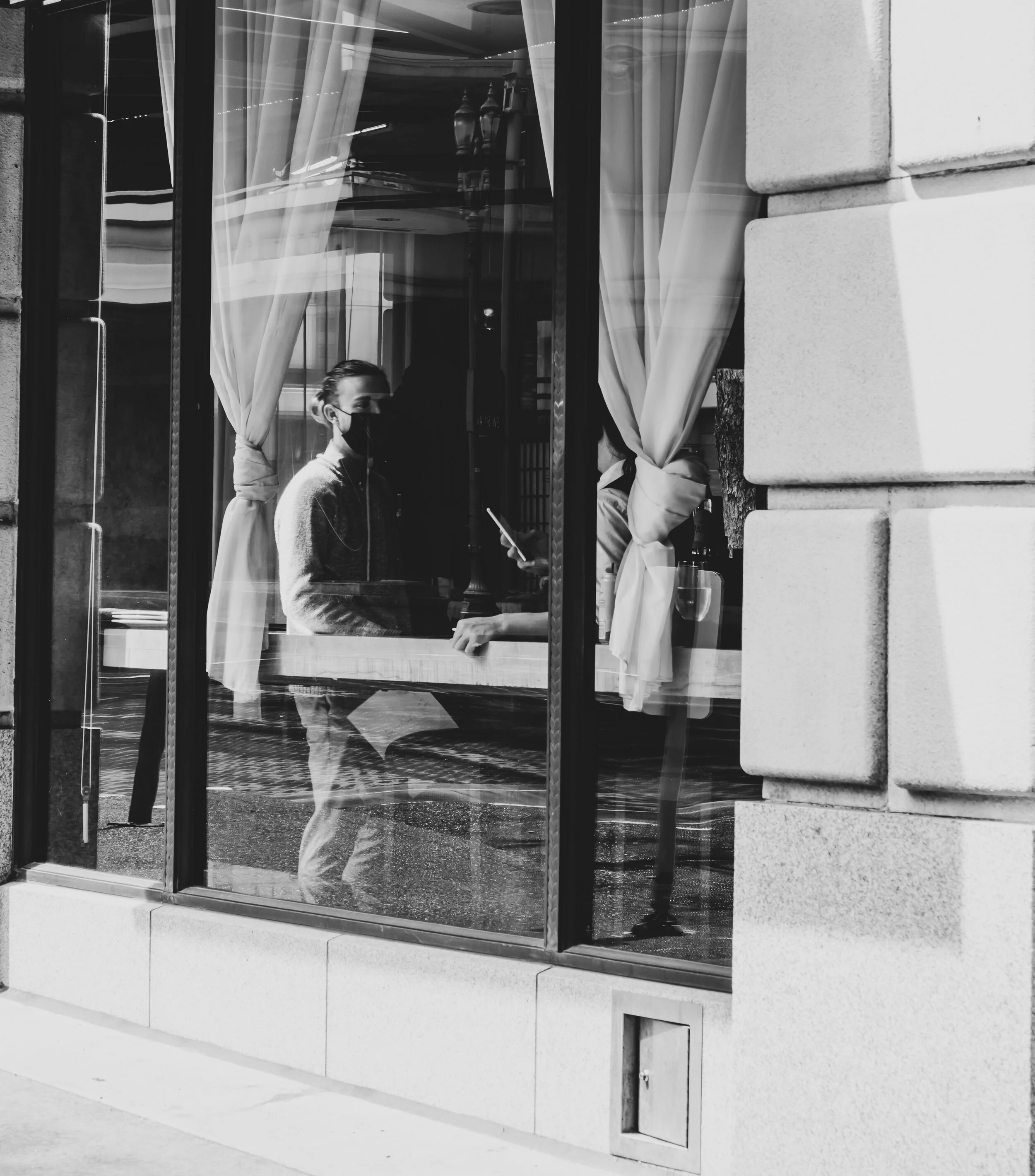 A person wearing a face mask inside a cafe, looking at someone across the counter, seen through a glass window with curtains.