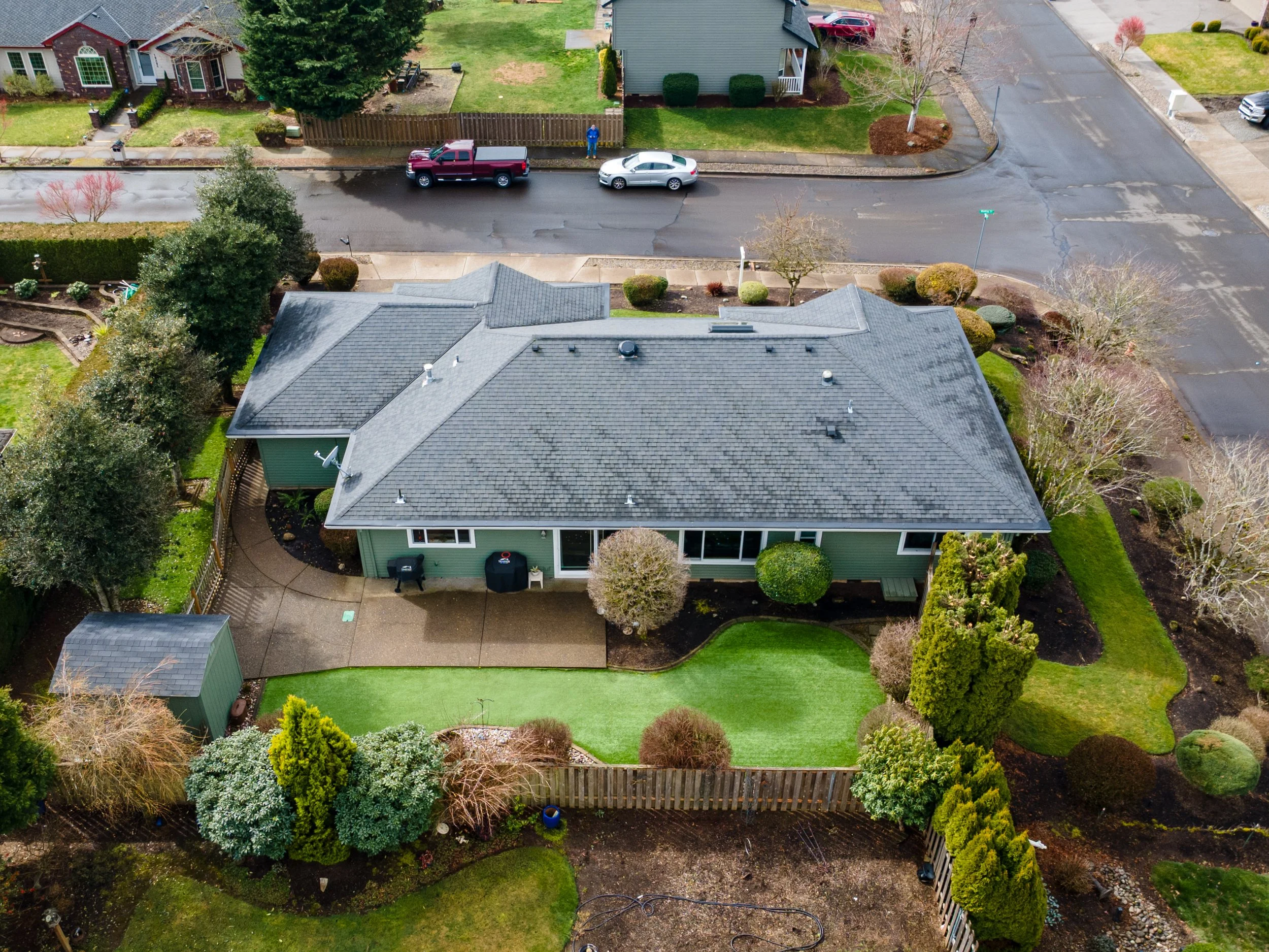 Aerial view of a suburban house with a gray roof and green exterior, surrounded by a landscaped yard with grass, shrubs, and trees, and a driveway with cars parked on the street.