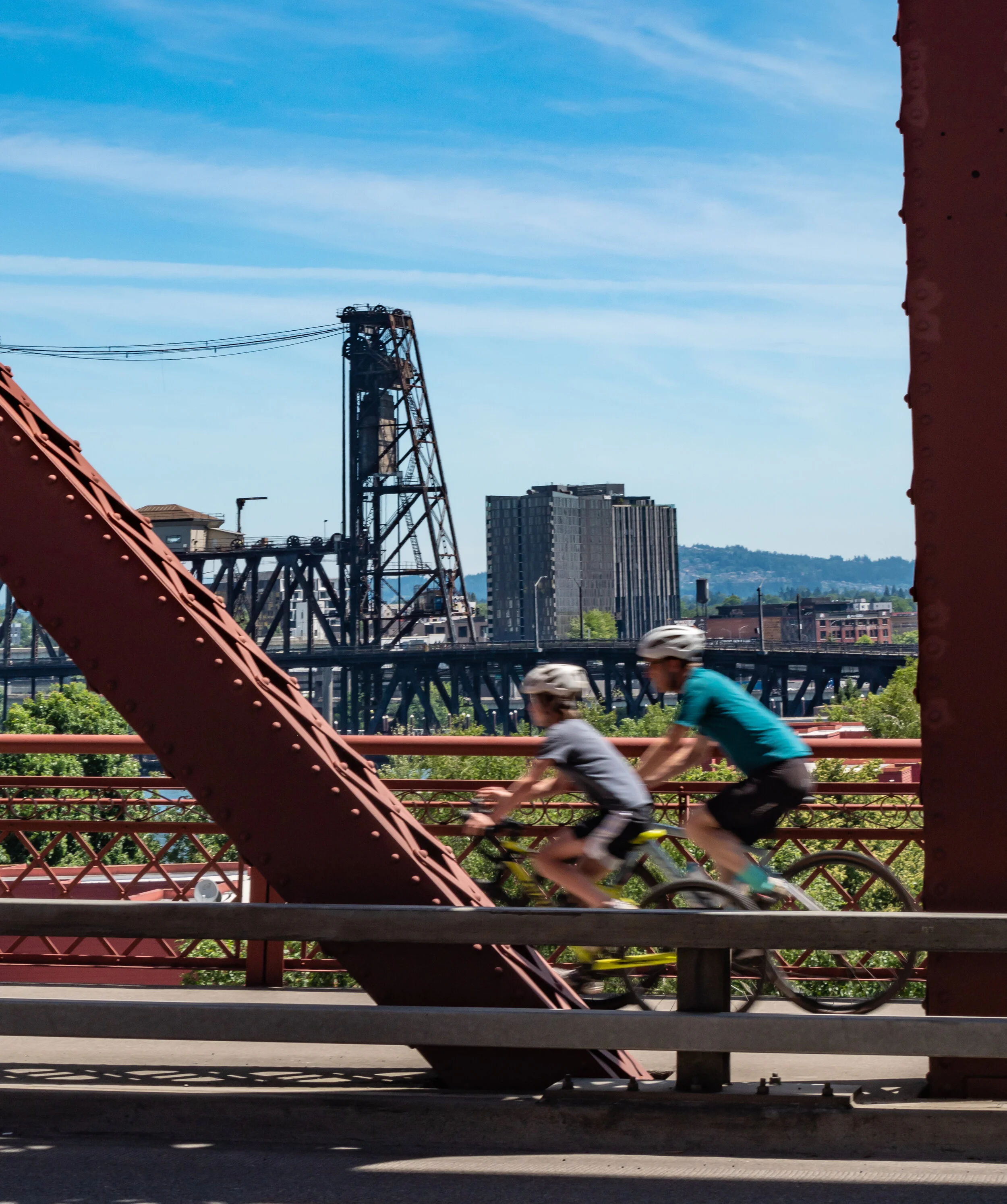 Two children riding bicycles on a bridge with industrial structures, buildings, and trees in the background under a blue sky.