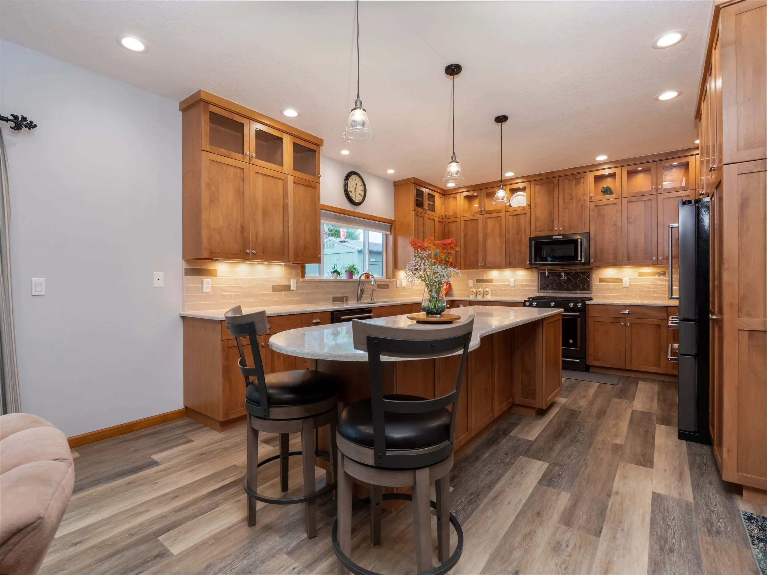 Kitchen with wooden cabinets, a farmhouse sink, black appliances, a kitchen island with seating, pendant lights, and wood flooring.