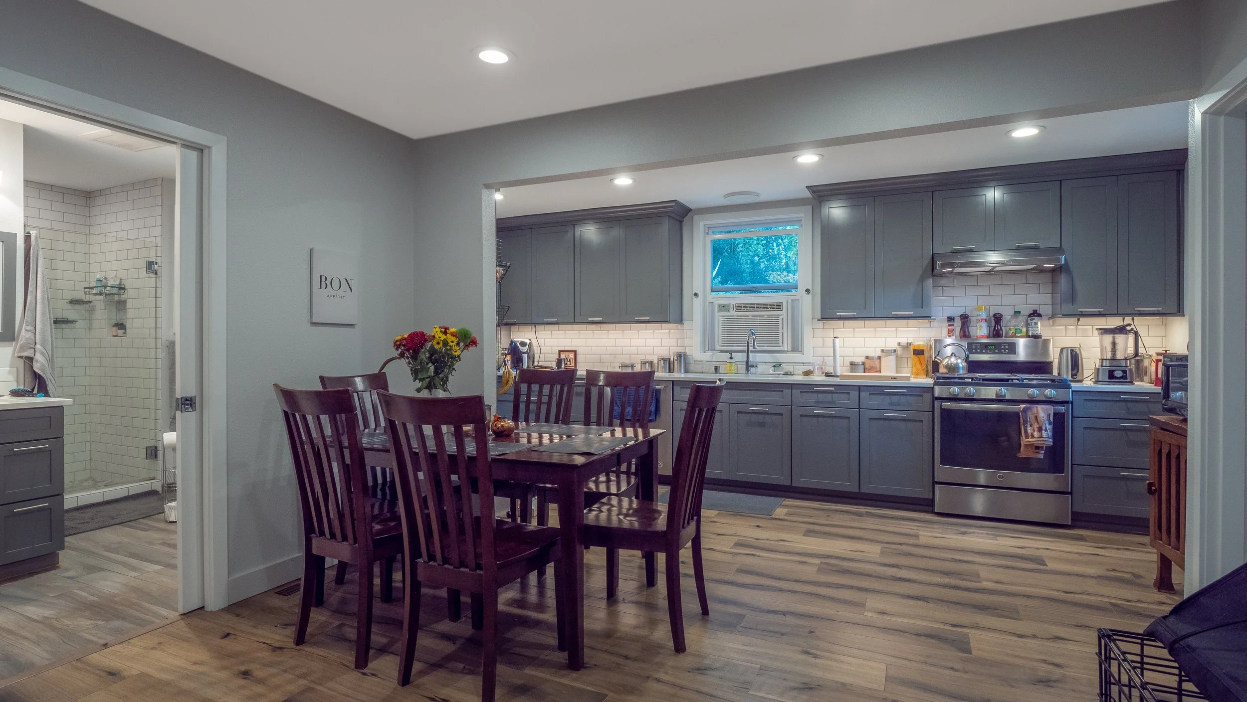 A modern kitchen with gray cabinets, stainless steel appliances, a dining table with six chairs, and a window air conditioning unit above the sink. To the left, there's a door leading to a bathroom or shower area.