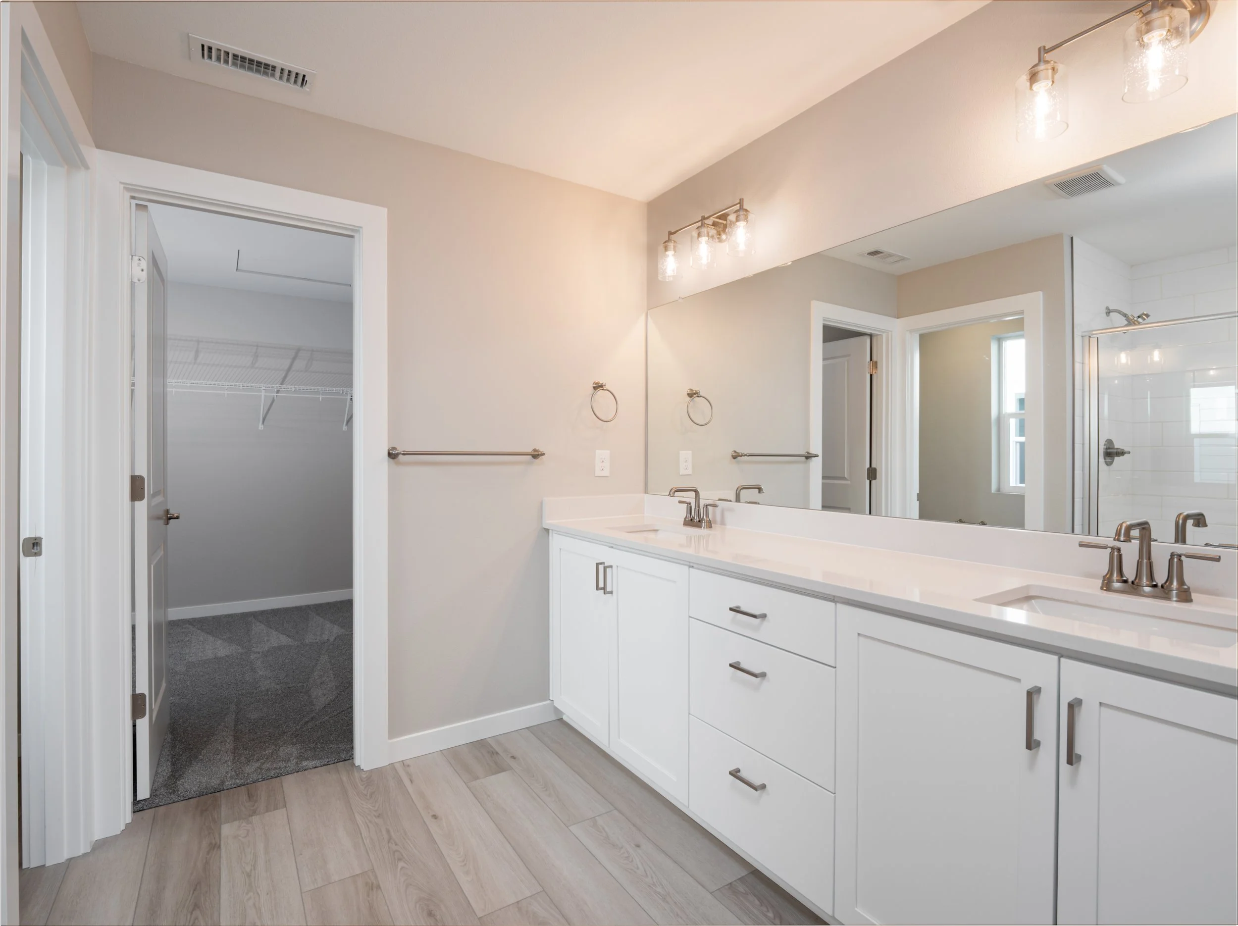 Modern bathroom with dual sinks, large mirror, beige walls, and a walk-in closet with shelving in the background.