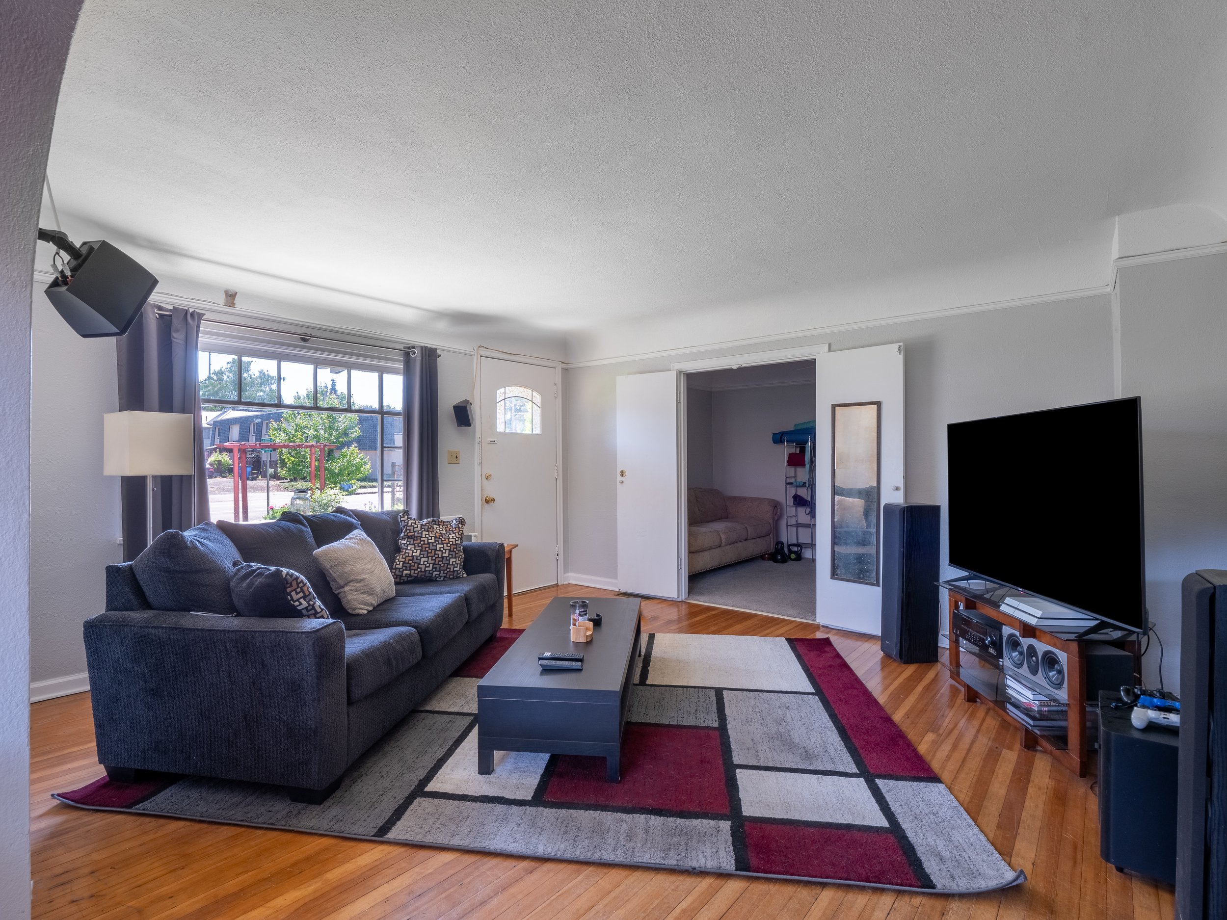 Living room with a blue sofa, coffee table, large window with gray curtains, TV on a stand, and various electronic devices. There's an open door to a small extra room with a beige couch and a mirror on the door. The room has hardwood floors, a patter