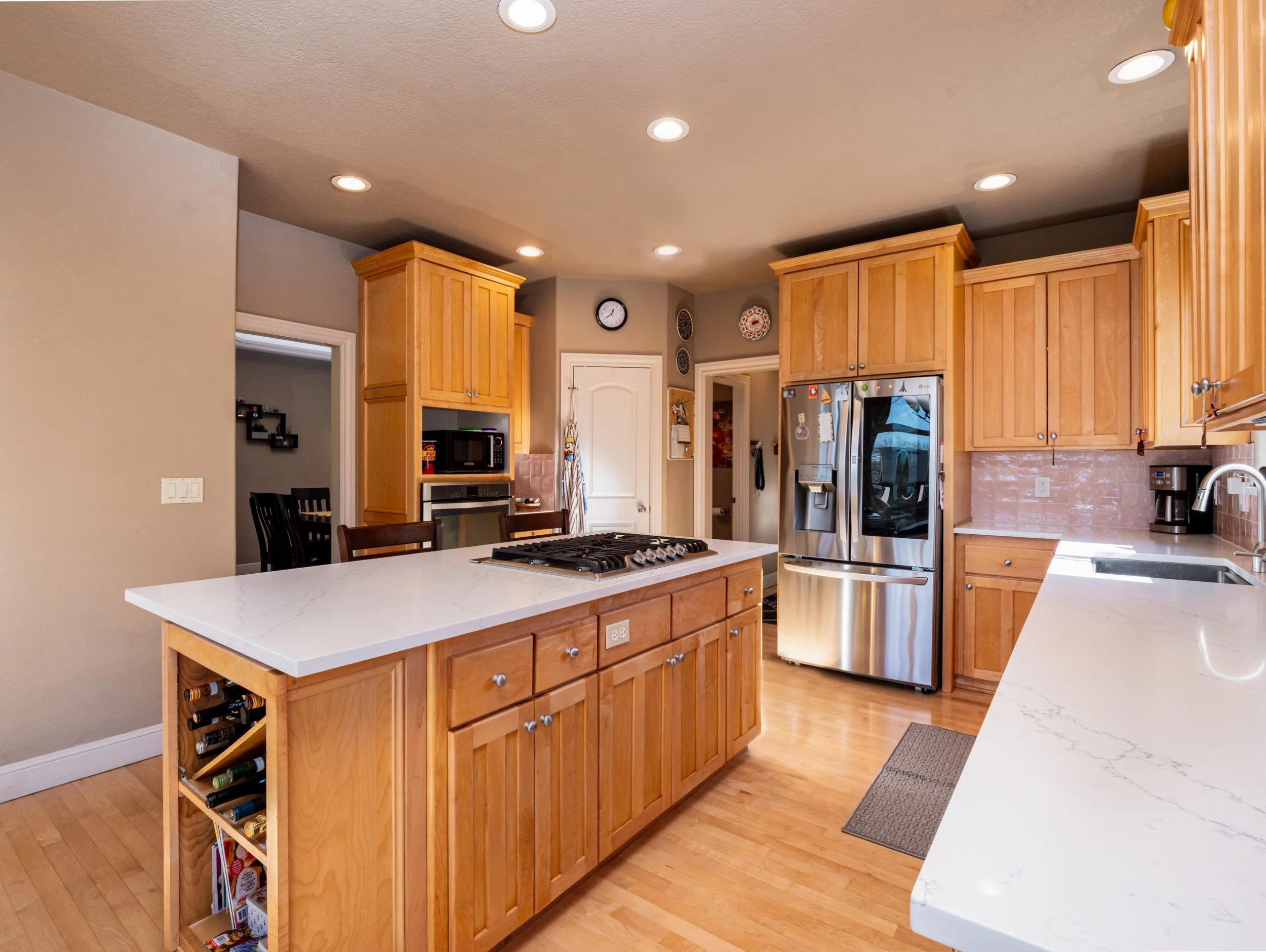 Modern kitchen with wooden cabinets, white marble countertops, stainless steel appliances including a refrigerator, microwave, and oven, and an island with a gas stove and wine storage. Recessed lighting and hardwood floors complement the space.