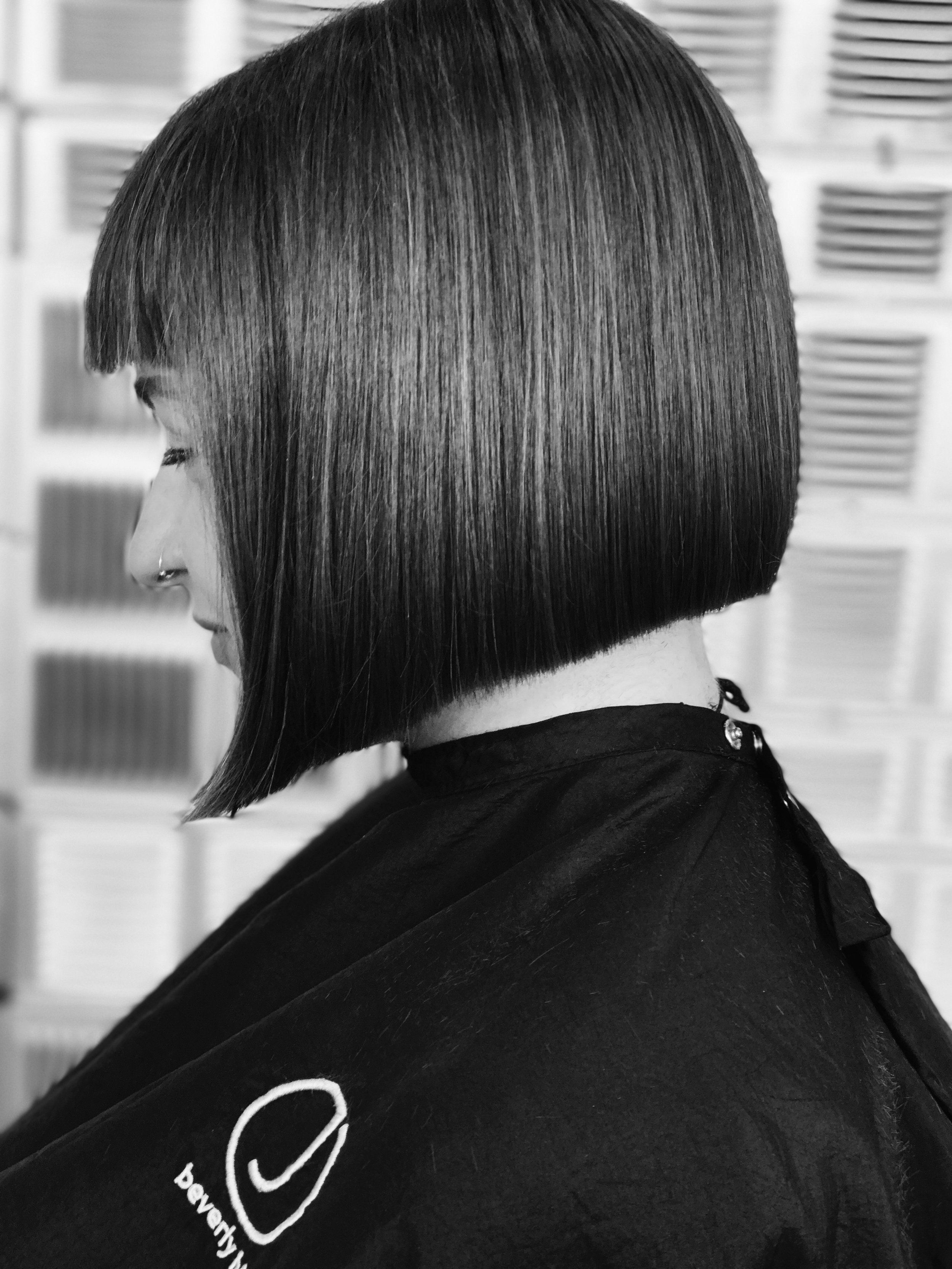 Black and white side profile of a woman with a sleek bob haircut and bangs, wearing a salon cape.