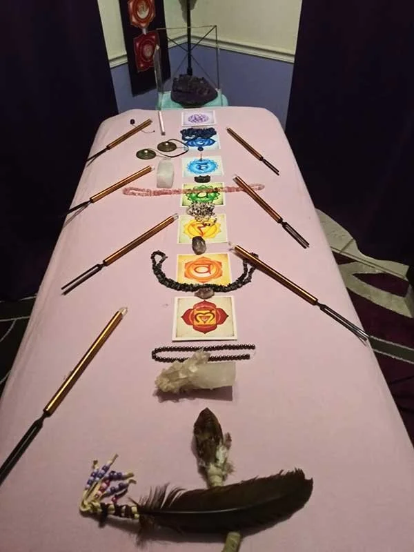 A table with various spiritual or ritual objects including candles, crystals, a feather, and symbolic cards, set in a room with dark curtains and artwork on the walls.