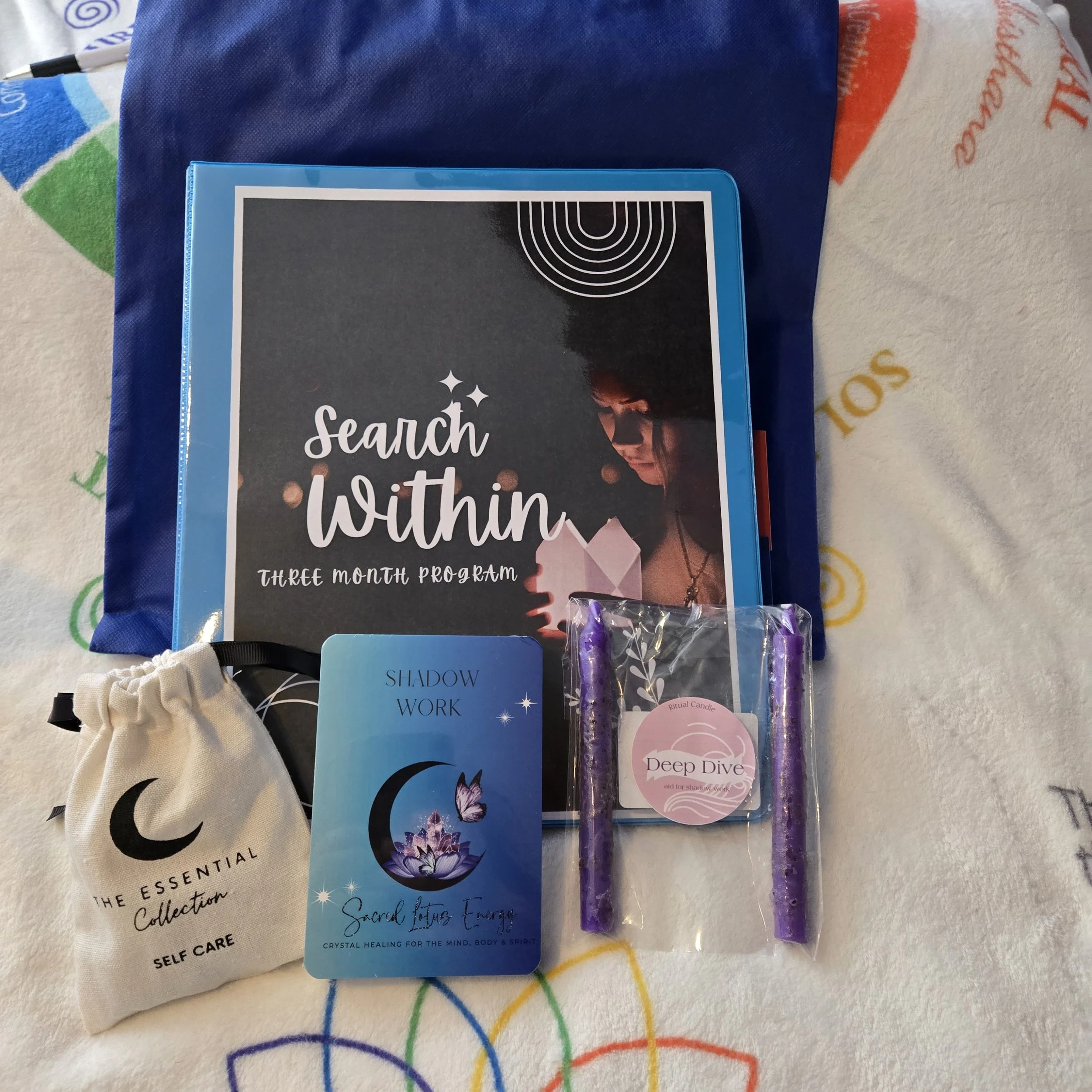 A variety of spiritual and self-care items including a book titled 'Search Within,' a shadow work card with a moon and butterfly design, a small bag labeled 'The Essential Collection Self Care,' a purple ritual candle called 'Deep Dive,' and a three-month program called 'Search Within' on a blue folder, all placed on a colorful patterned fabric.