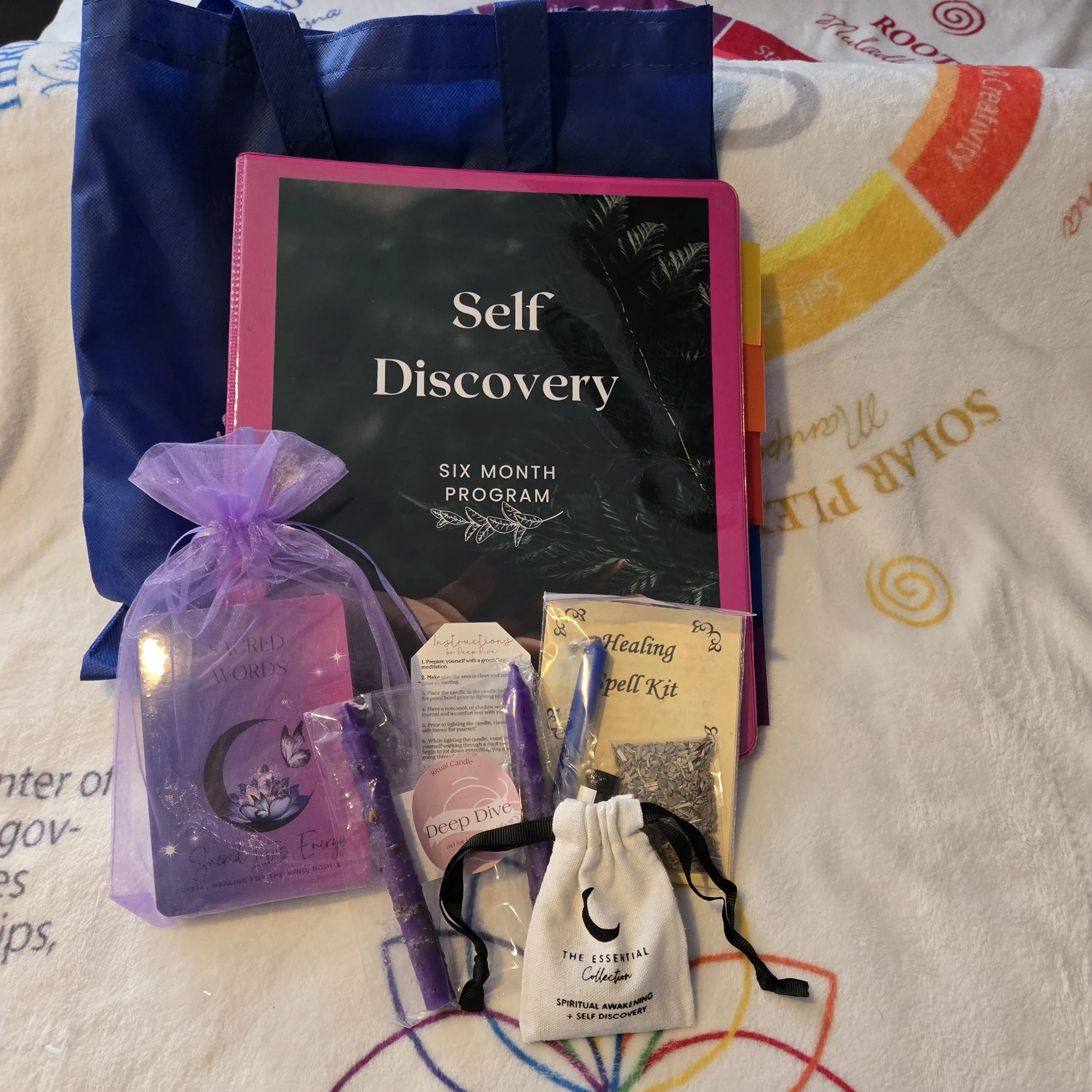 A self-discovery six-month program kit includes a large black and pink binder, a purple moon-themed gift bag with crystals and incense, a healing spell kit, a small pouch labeled 'The Essential Collection,' and a blue tote bag, arranged on a colorful cloth.