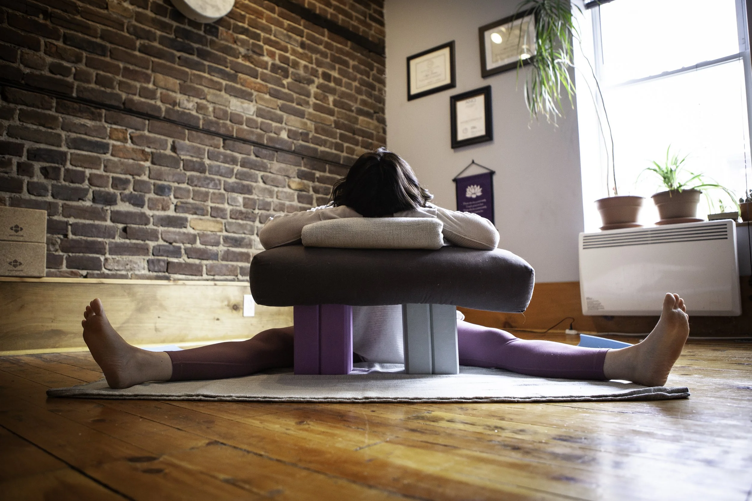A woman practicing yoga on a purple yoga mat in a room with an exposed brick wall, framed certificates, and two potted plants near a window.