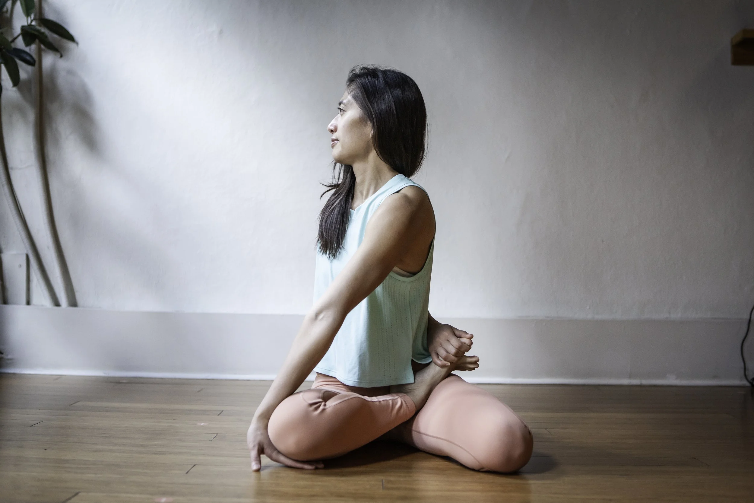 A woman practicing yoga indoors on a wooden floor, seated in a cross-legged position while holding one foot behind her back. She is dressed in a light blue tank top and light pink leggings, with long dark hair and a focused expression. The background features a plain white wall and some plant leaves on the left side.