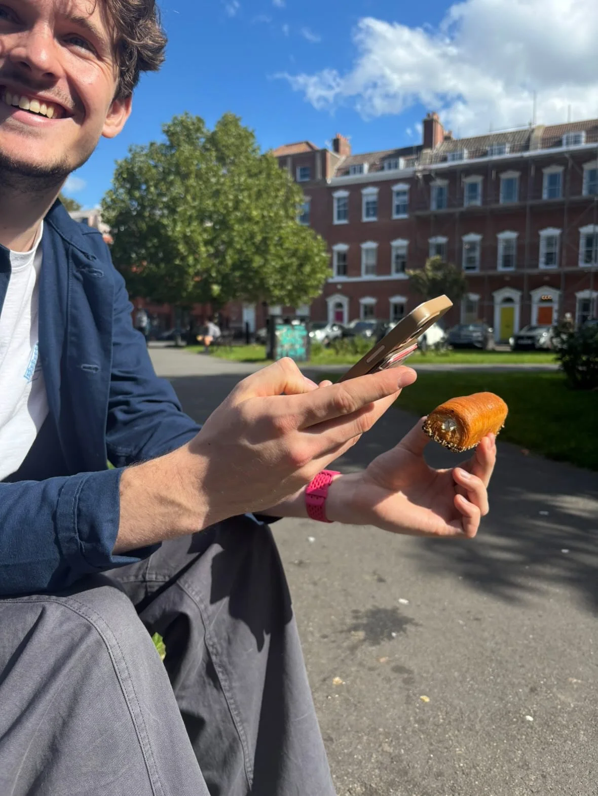 A smiling man holding a smartphone in one hand and a partially eaten pastry in the other hand, sitting outdoors on a sunny day with trees and brick buildings in the background.