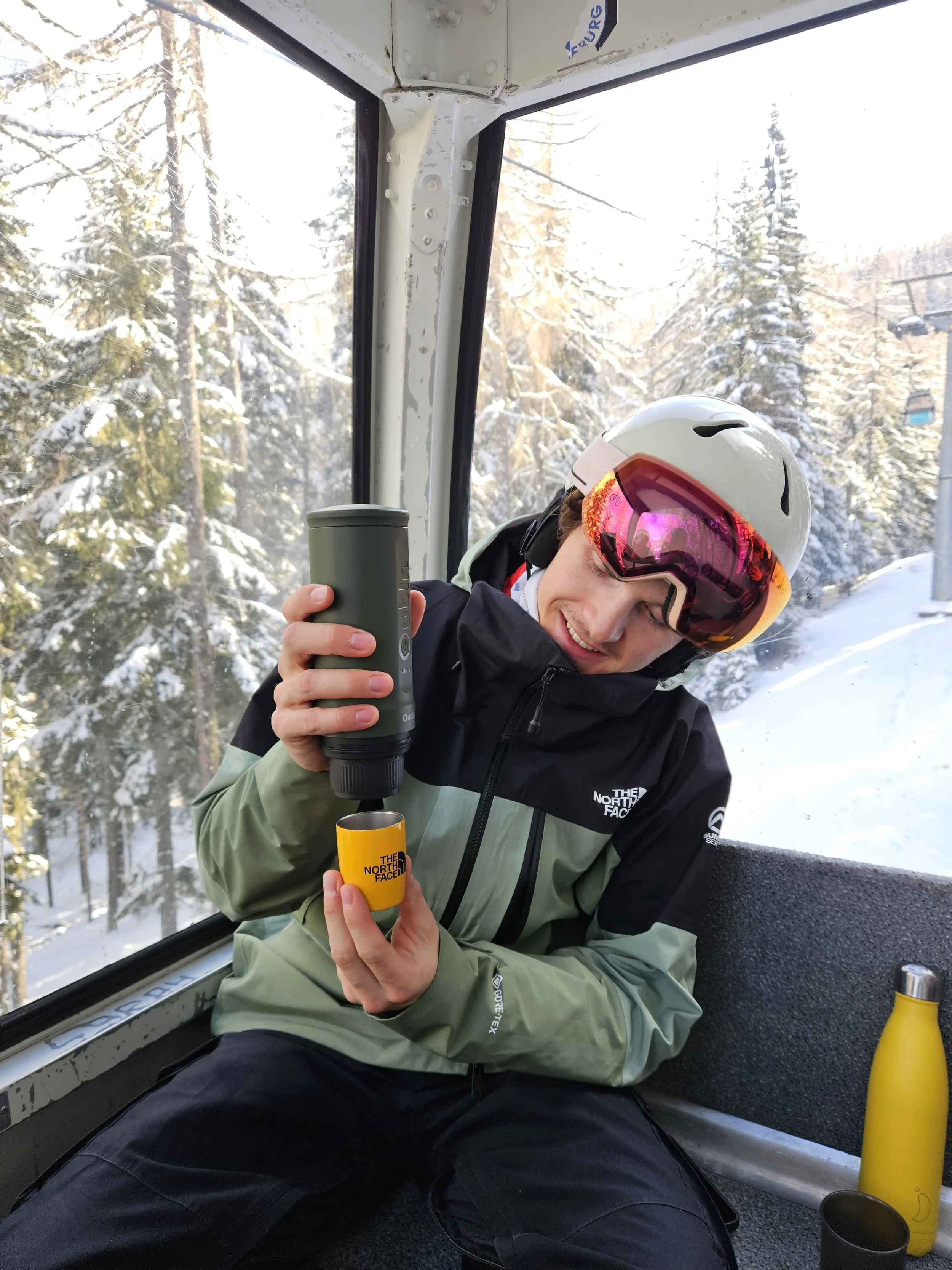 A person in winter gear, including a white helmet with a pink visor and a green North Face jacket, is smiling and pouring a drink from a thermos into a yellow cup that has the North Face logo. The person is sitting inside a ski lift cabin with snowy trees outside the window.