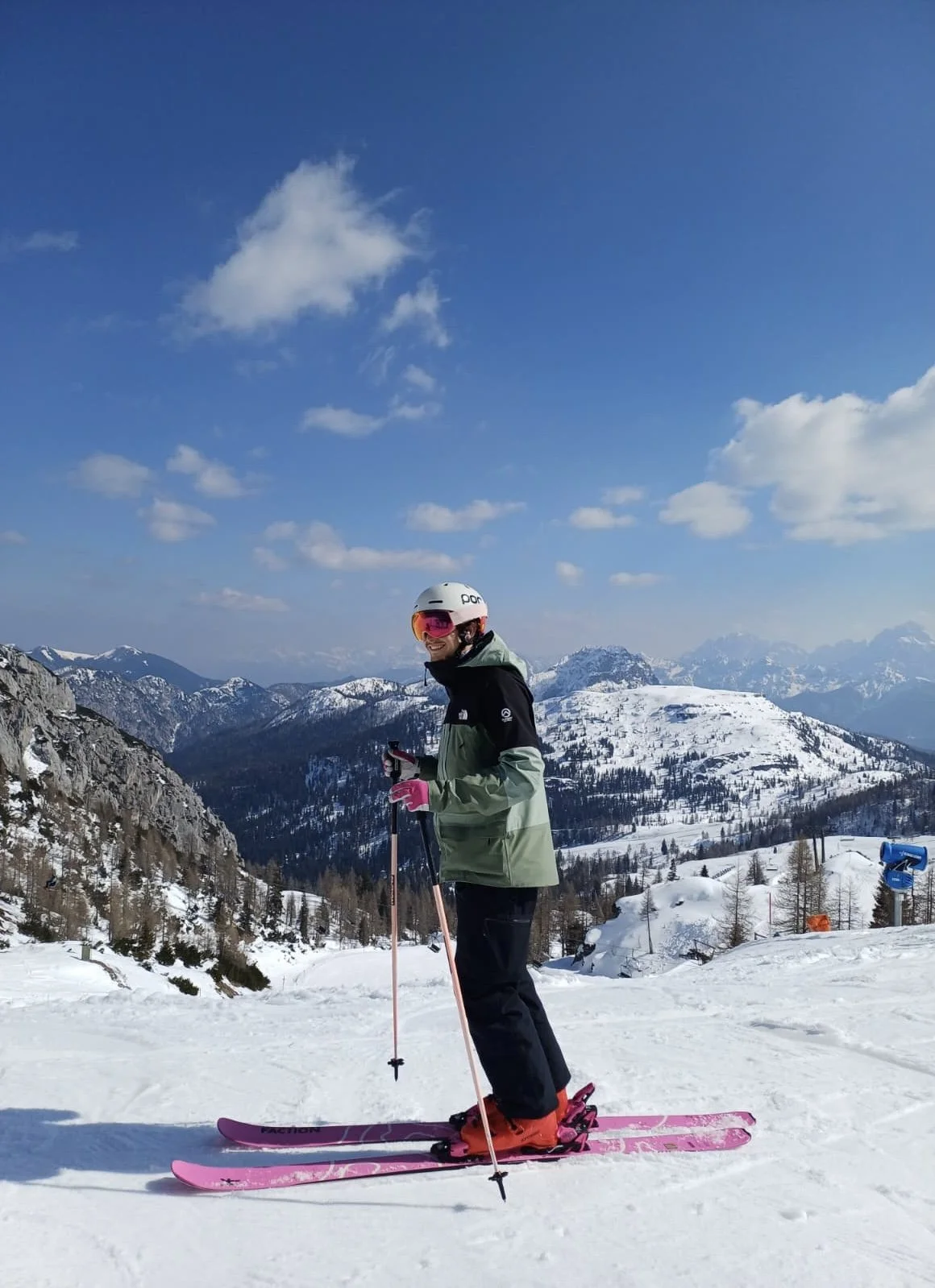 Person in ski gear, smiling and holding ski poles, standing on a snow-covered mountain slope with snow-capped mountains in the background under a partly cloudy blue sky.