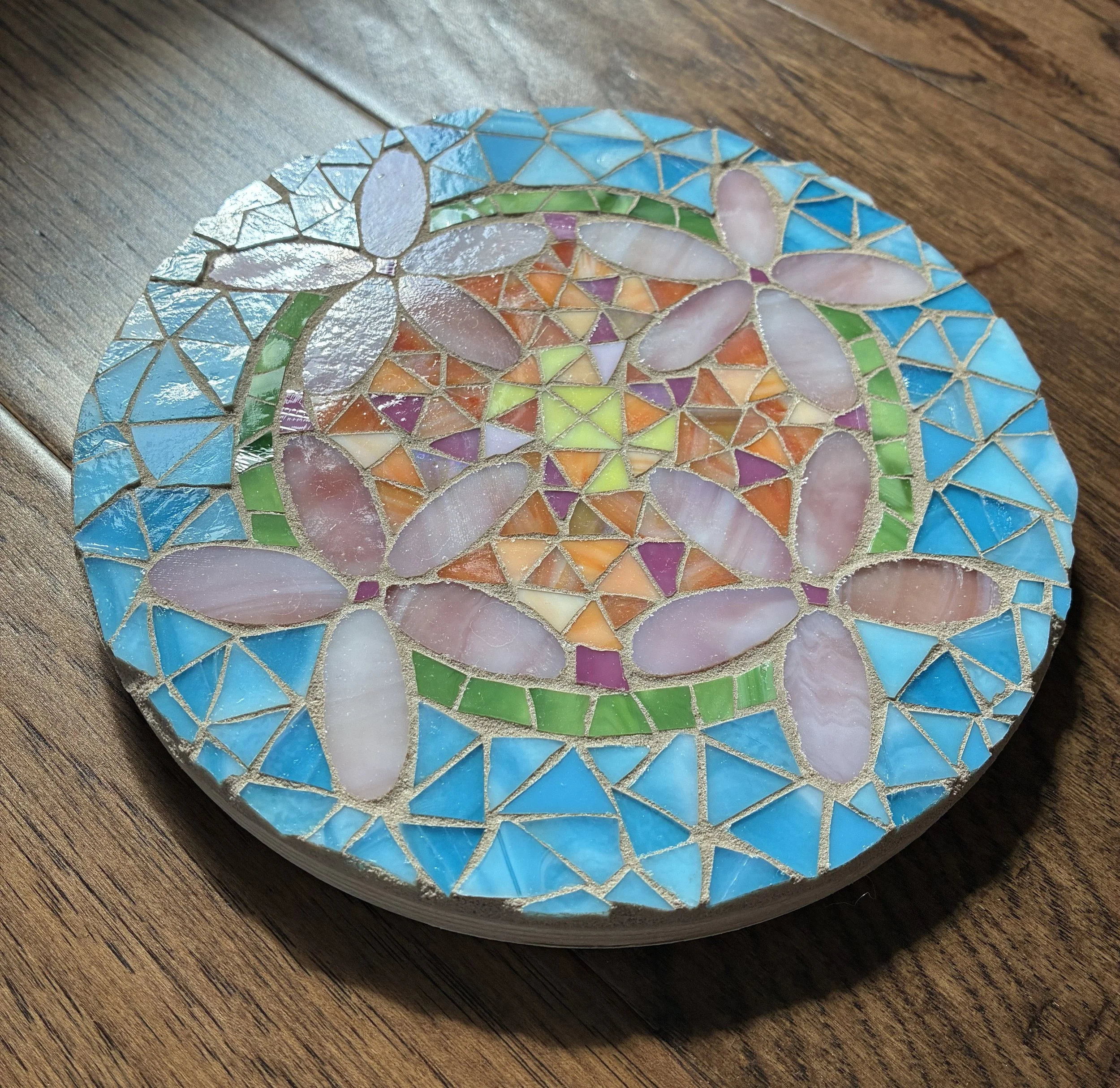 A round mosaic art piece with floral and geometric patterns, made of colorful glass pieces, placed on a wooden surface.