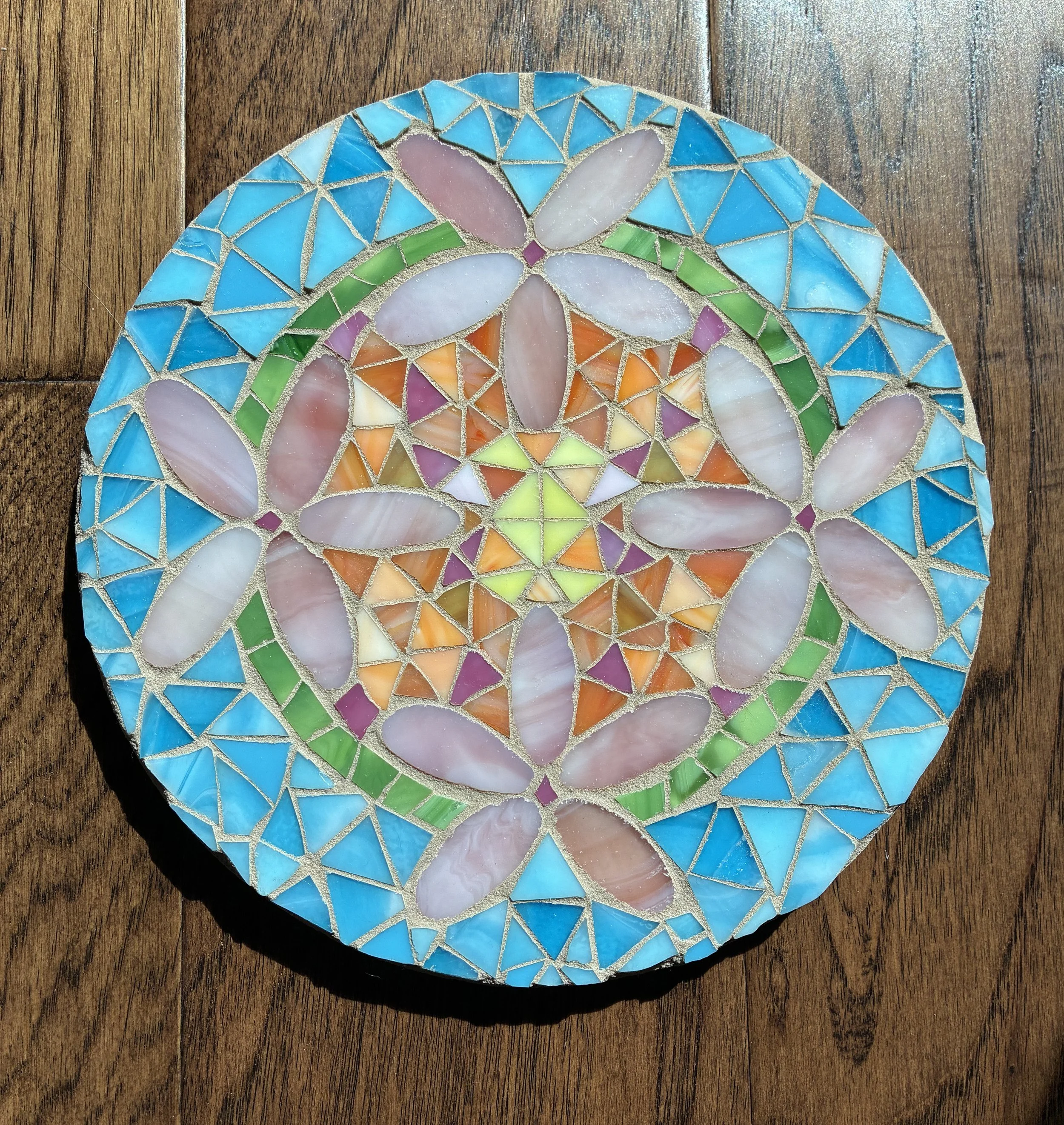 Mosaic art featuring a butterfly design in colors of pink, cream, orange, purple, and green on a round, blue background, placed on a wooden surface.