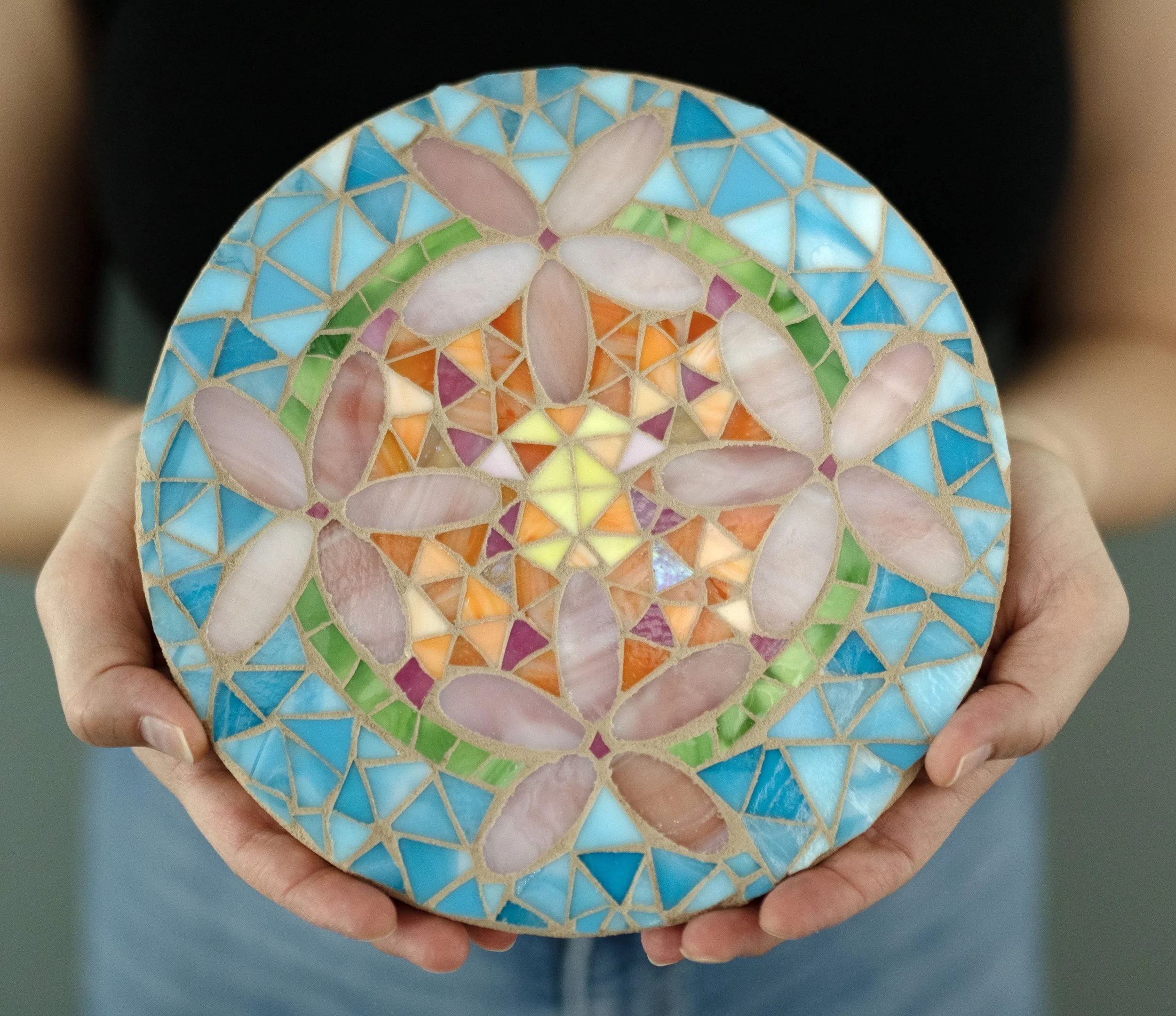 Close-up of a hand holding a circular mosaic artwork featuring a butterfly design with pastel colors, including pink, purple, yellow, orange, green, and blue, set against a blurred background.