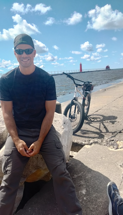 Man in black shirt wearing sunglasses and backwards black hat sits on a rock with a bike and lighthouse in the background