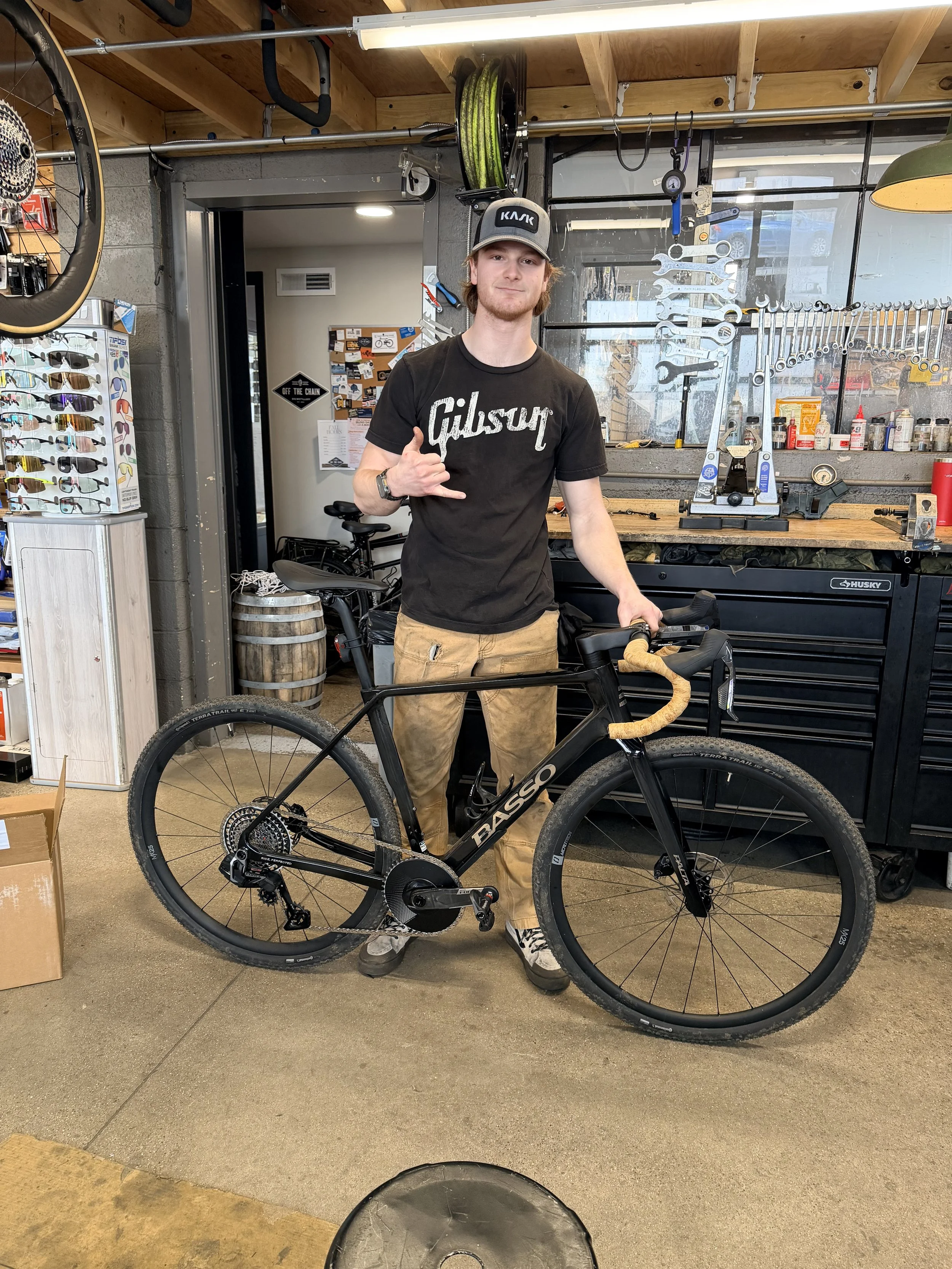 Young man in black shirt, khaki pants, and a hat stands behind black road bike in bike shop