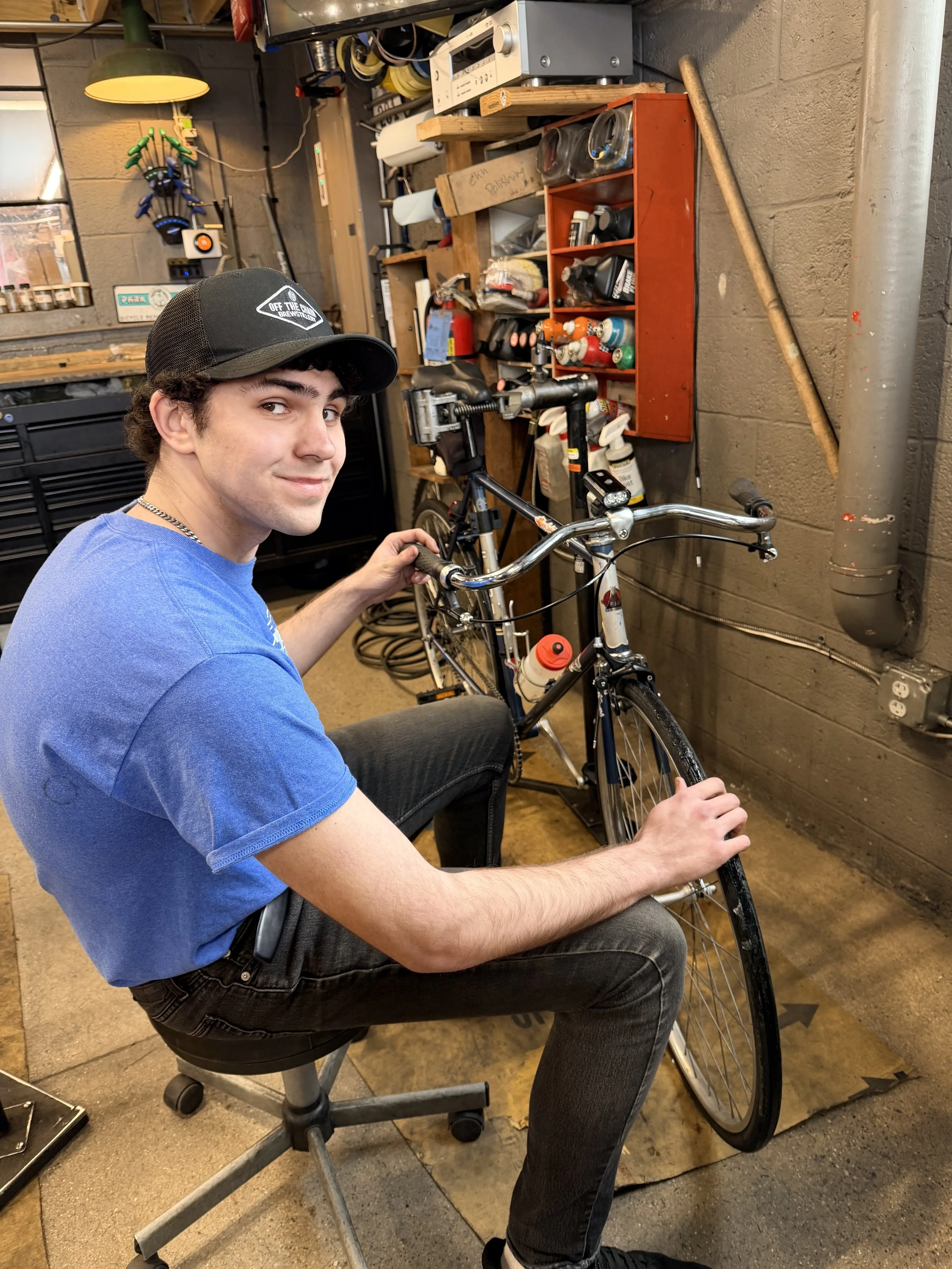 Young man in blue shirt and black hat sits in front of bike in stand while working on the bicycle