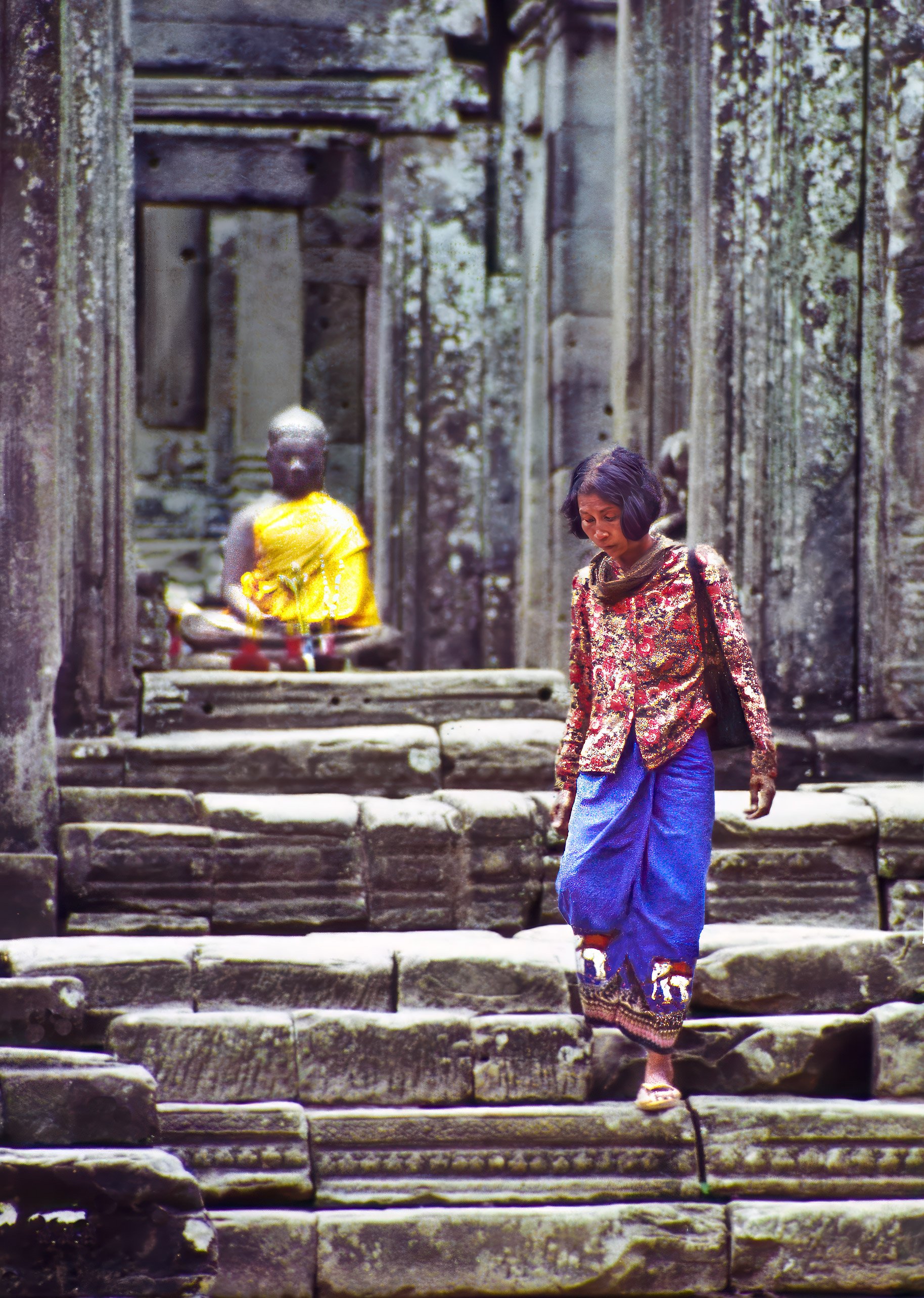 A woman descends the steps of an Ankor Wat temple under the inanimate gaze of a statue of the Buddha.