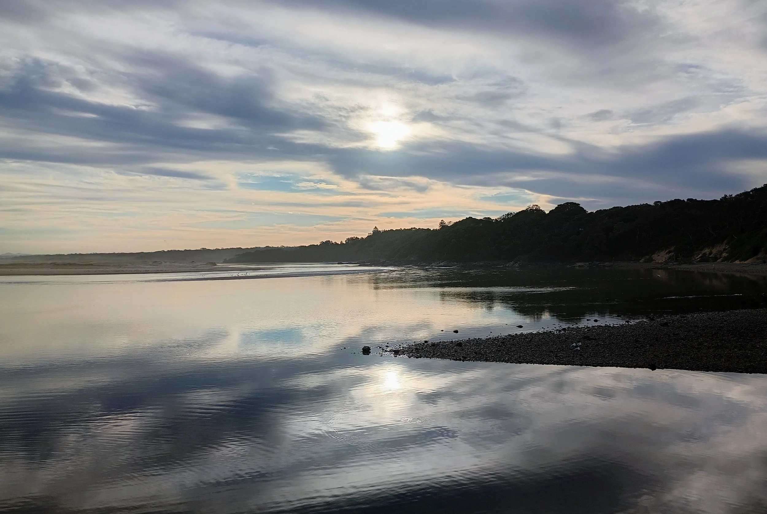 Sky with clouds over a calm body of water reflecting the sky, with a distant tree-covered shoreline.