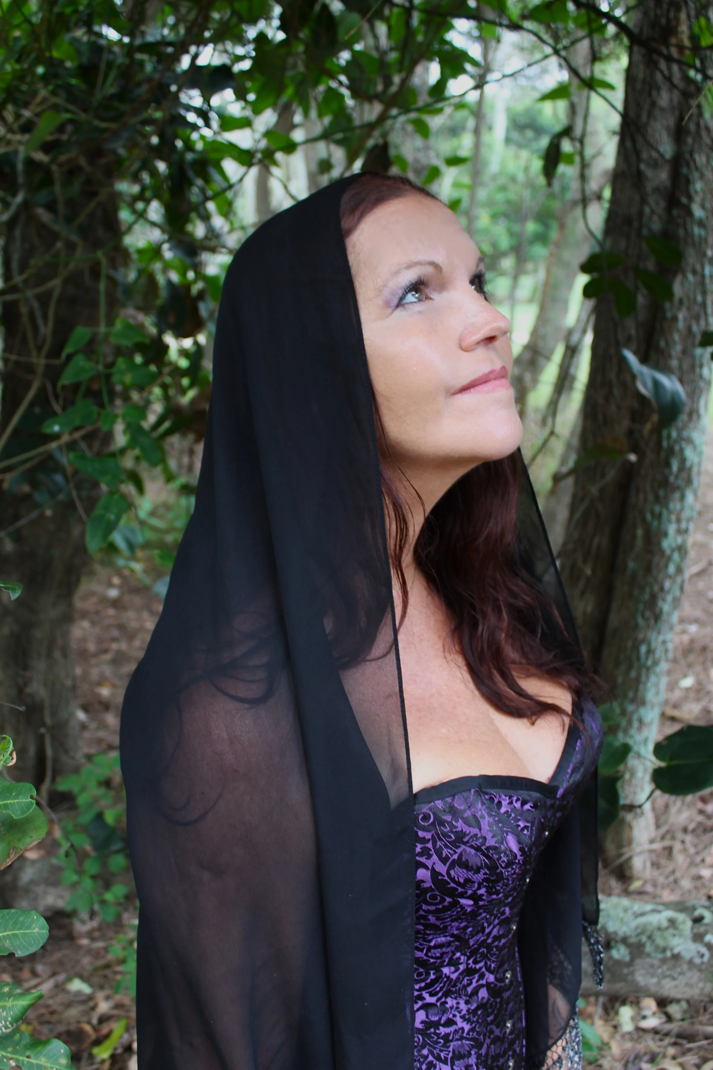 A woman with dark red hair and light skin wearing purple and black lingerie and a black sheer shawl, standing outdoors among trees and green foliage, looking upward.