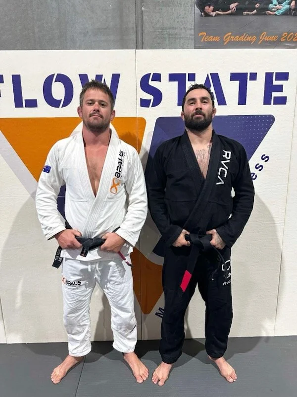 Two men standing in Brazilian Jiu-Jitsu gi uniforms on a mat against a wall. The man on the left wears a white gi, the man on the right wears a black gi. Both are barefoot and holding their black belts in front of them.