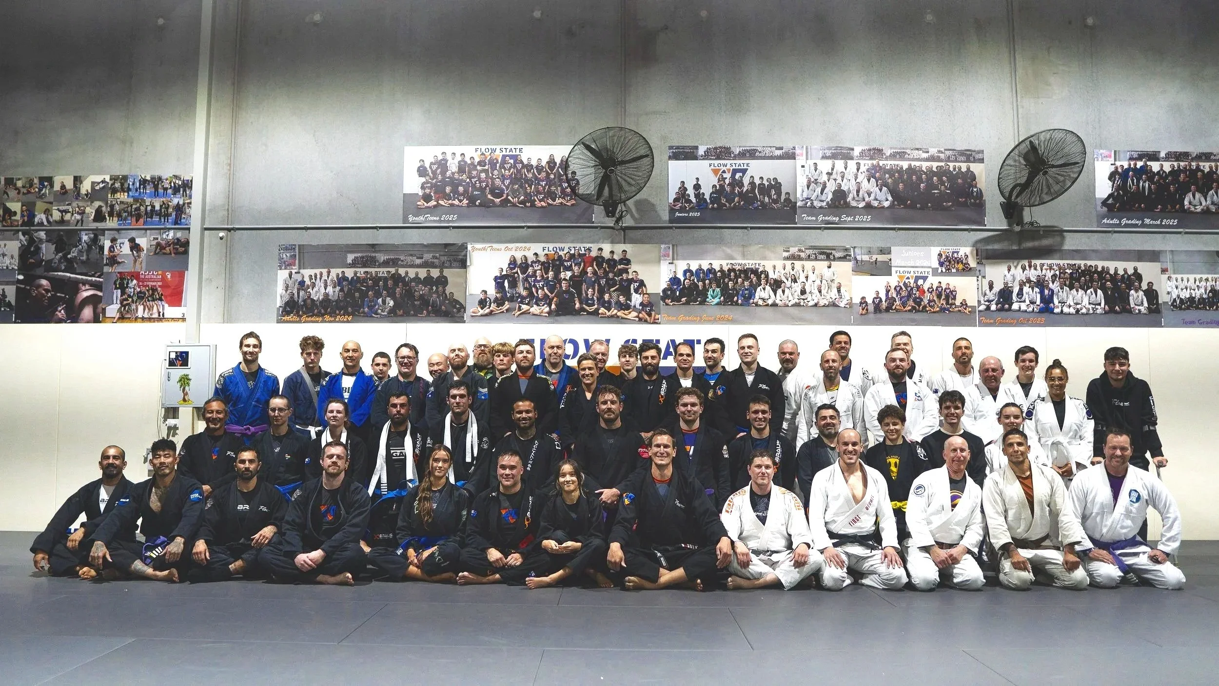 Group photograph of martial artists in uniform at a dojo, with banners displaying team photos and event dates in the background.