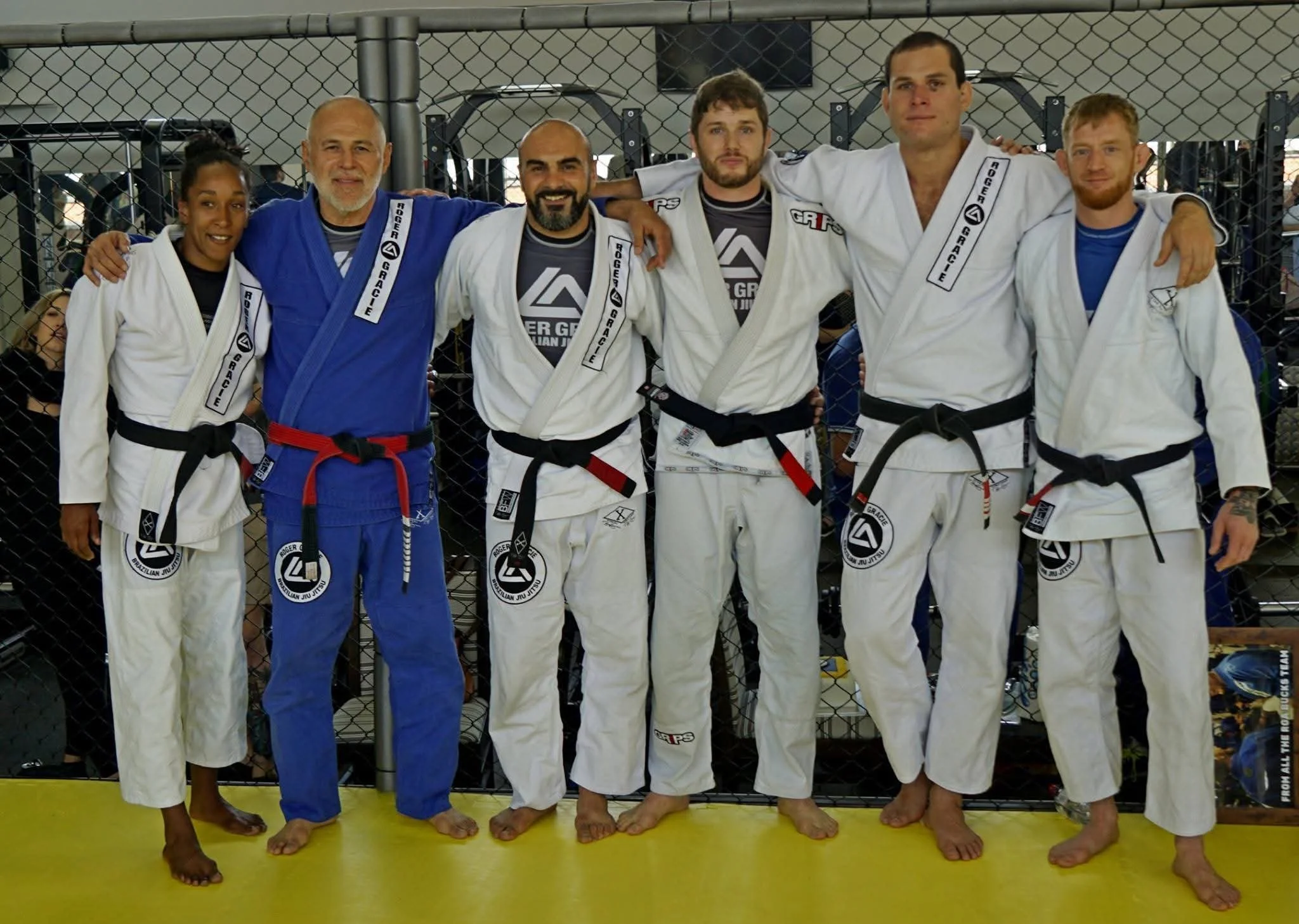 Group of six mixed martial artists and fighters standing together in a gym, all wearing gis with belts and patches, some smiling and some looking serious, behind a wire fence.