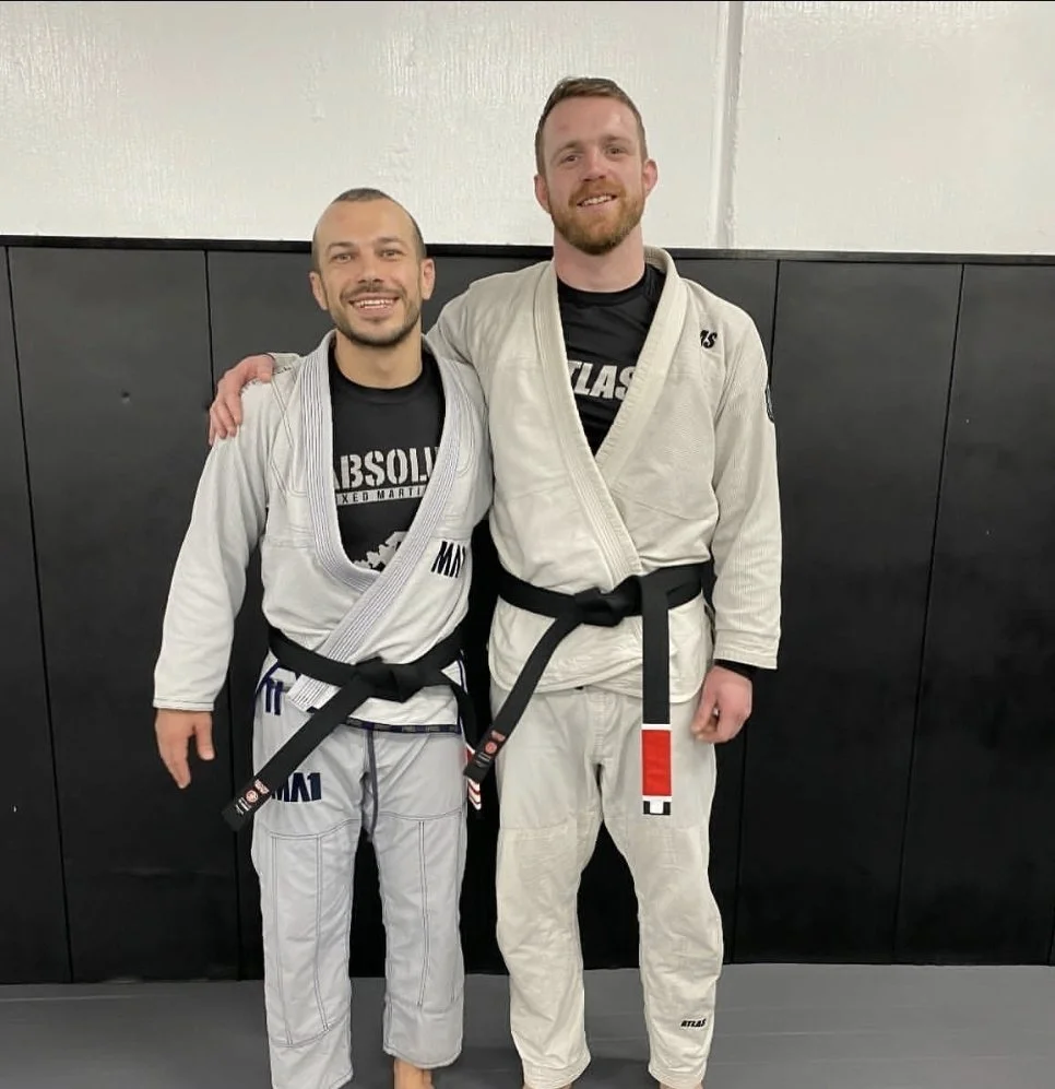 Two men in Brazilian Jiu-Jitsu gis, standing together and smiling, in a martial arts training facility. One man has his arm around the other's shoulder.