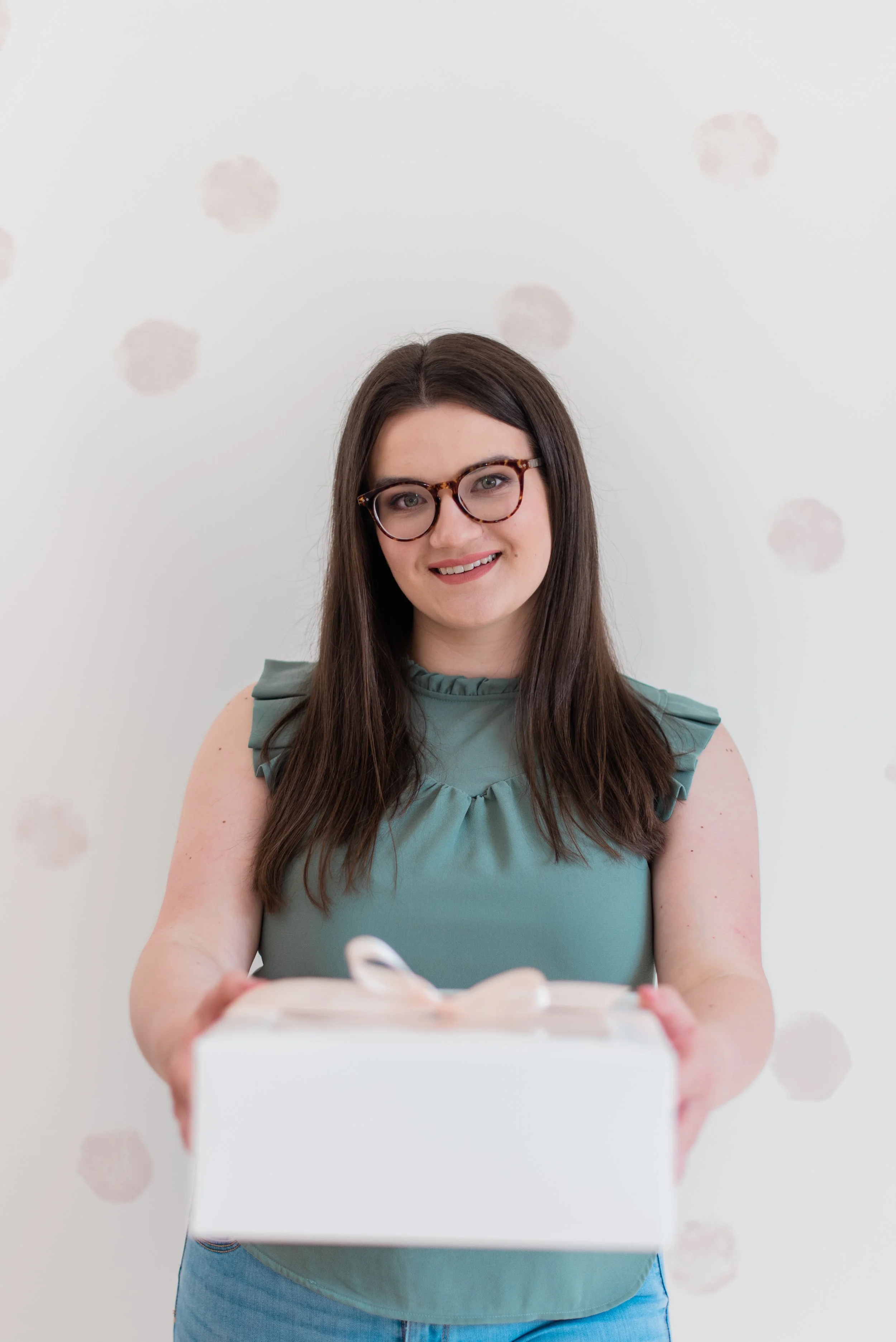 A woman with long dark hair and glasses smiling while holding a wrapped gift box with a ribbon.