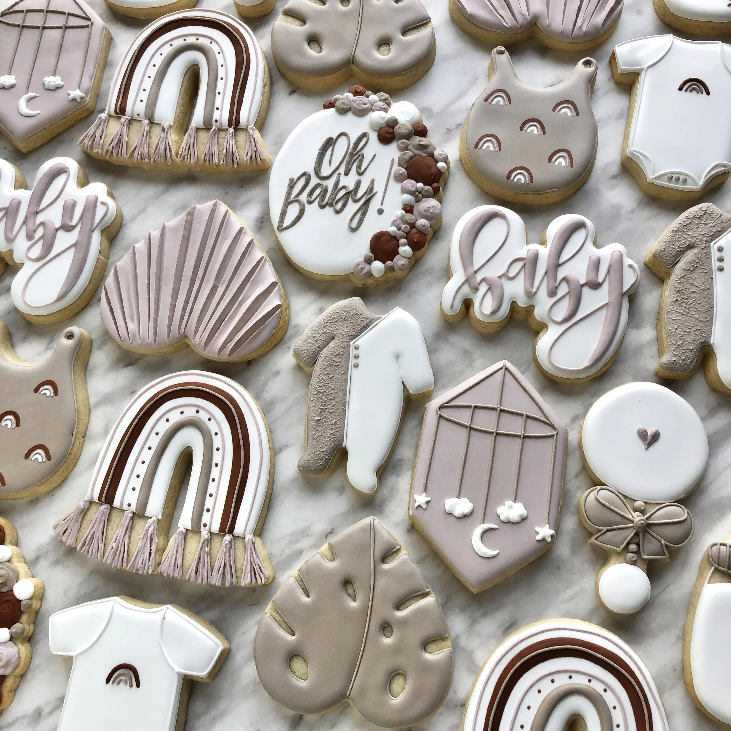 Custom decorated baby shower sugar cookies in pastel pink, brown, and white colors, with designs including rainbows, hearts, leaves, rainbows, baby onesies, and a hanging crib mobile with clouds, stars, and moon. Modern boho style party favours.