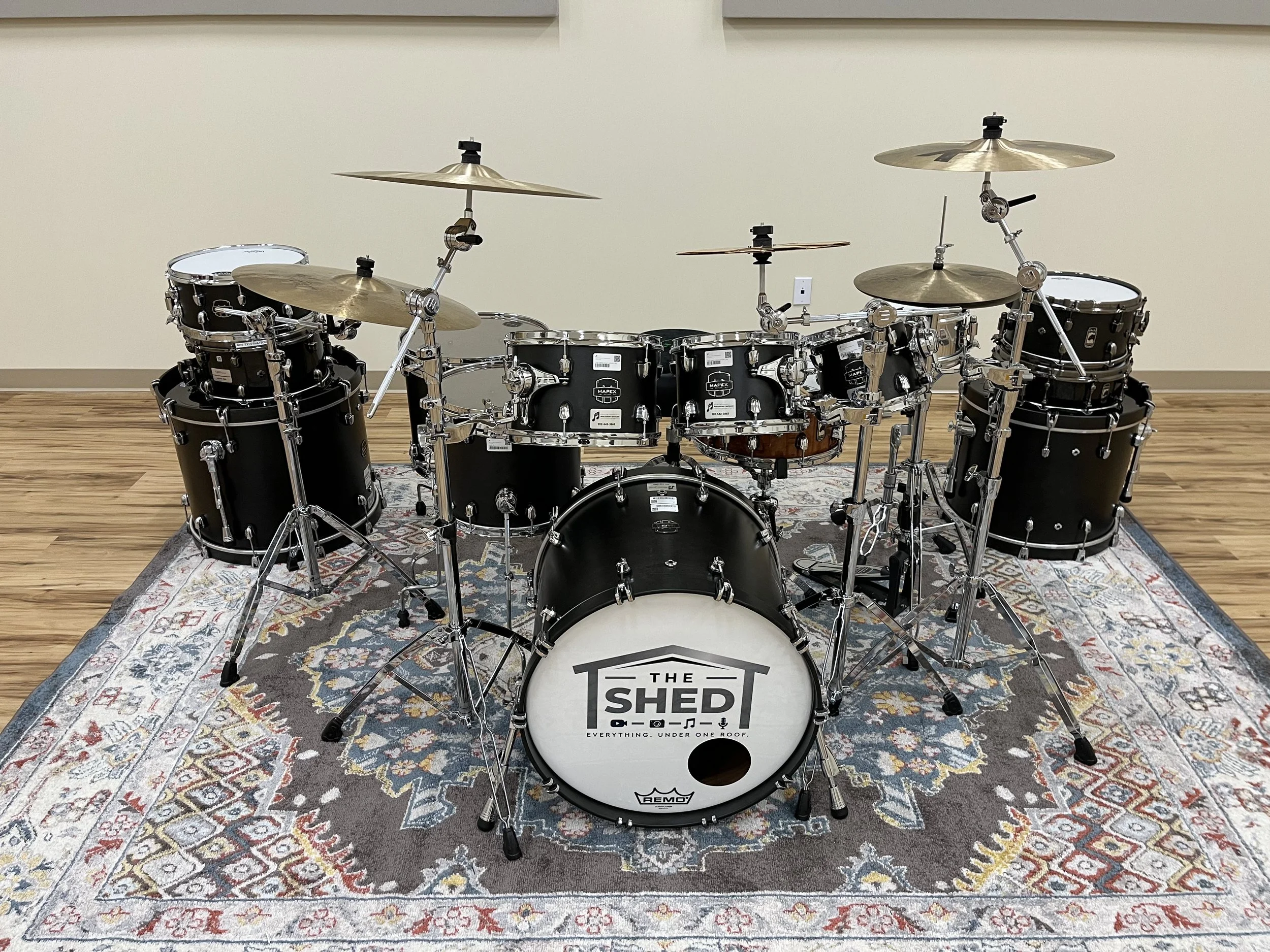A black drum set with silver hardware and cymbals arranged on a patterned rug in a room with wooden floors and a beige wall. The bass drum has a logo that reads 'The Shed.'