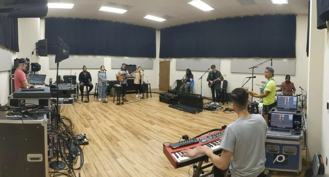 A music rehearsal room with musicians practicing, including a keyboard player, guitarists, vocalists, and a drummer, surrounded by audio equipment and soundproofing panels.
