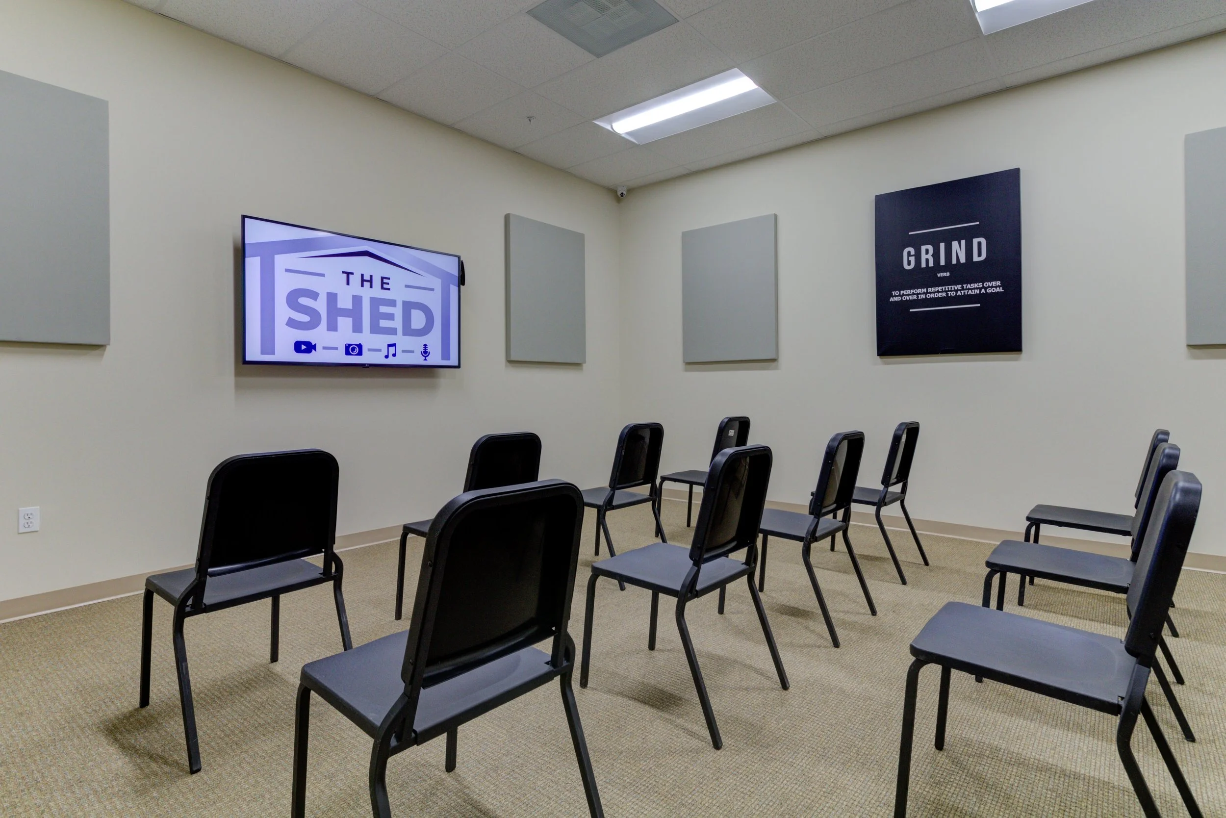 A small room with beige carpet, cream-colored walls, and a drop ceiling with fluorescent lights. There are multiple black chairs arranged in rows facing a wall with a mounted flat-screen monitor displaying 'The Shed.' Two black posters with motivatio
