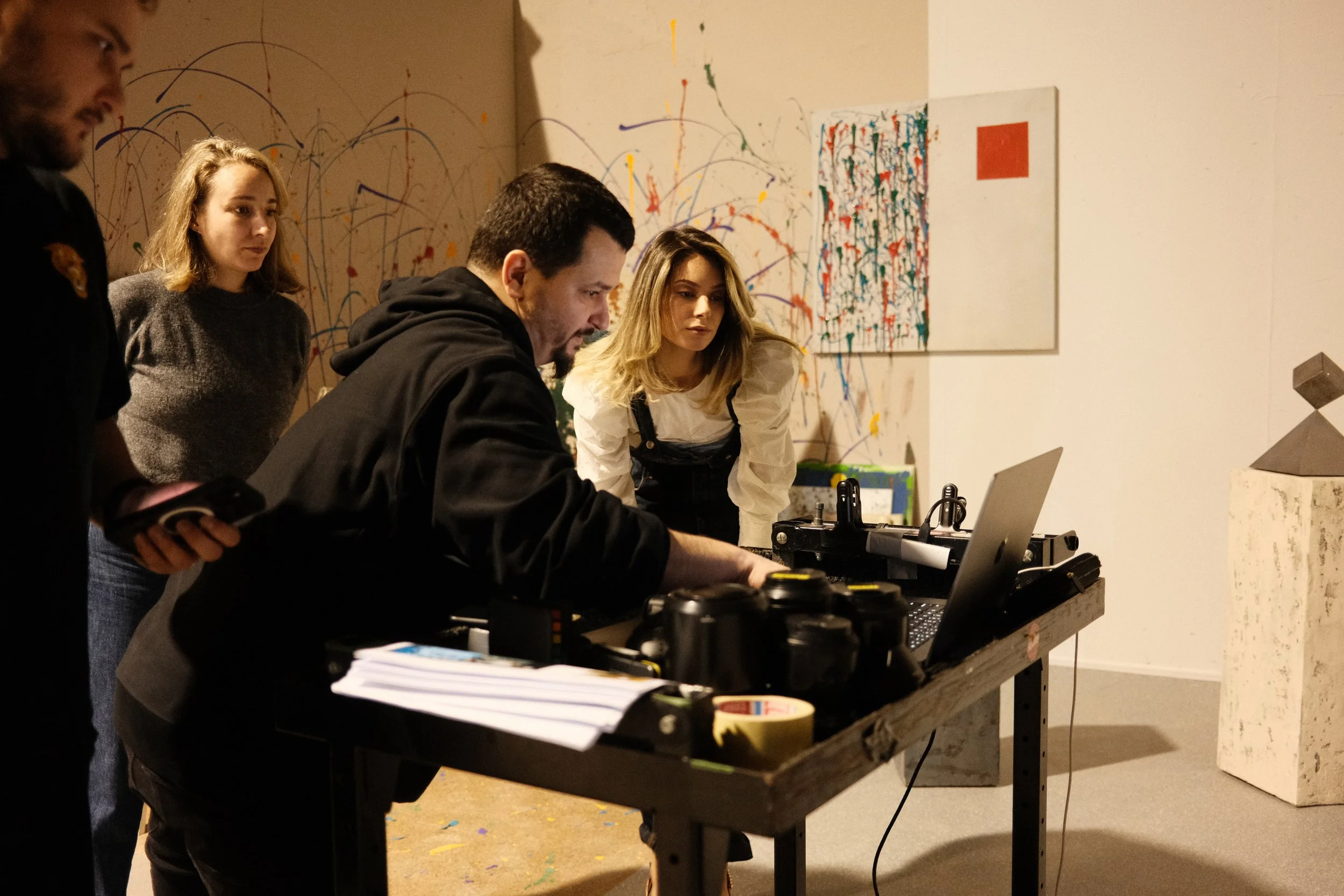 Four people are gathered around a table with DJ equipment in an art gallery. Behind them, there is a wall with colorful abstract paintings and paint splatters. The setting appears to be an art event or workshop.